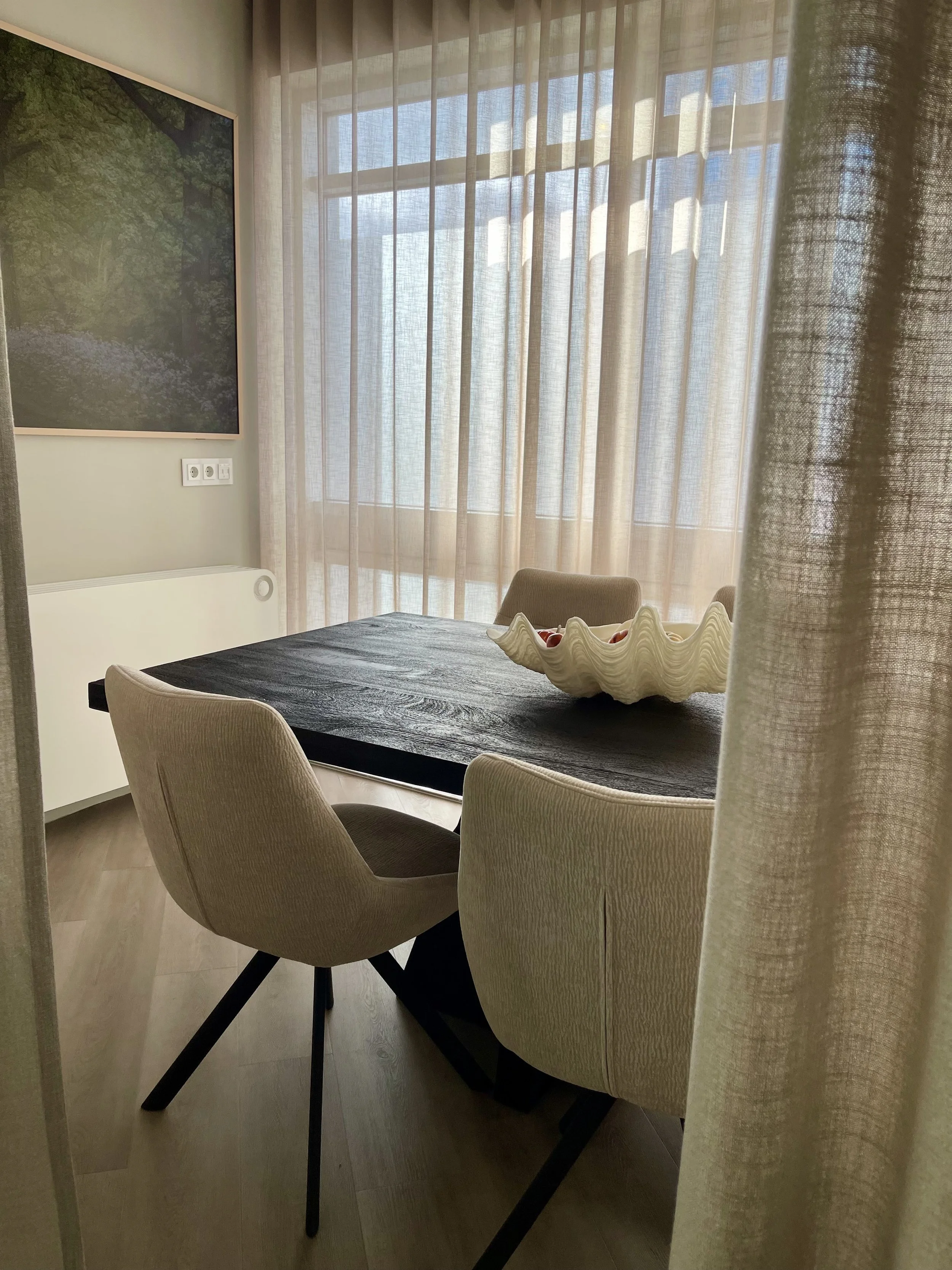 A dining room viewed through a curtain, featuring a dark wooden table with a ceramic bowl of fruit, beige upholstered chairs, a large window with sheer curtains, a framed art piece of greenery and water, an electrical outlet, and a white radiator.