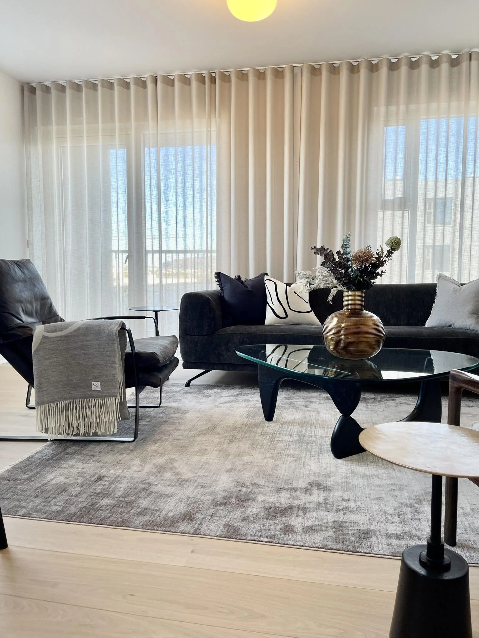 Living room with large windows covered by sheer curtains, a black sofa with decorative pillows, a glass coffee table with a vase of flowers, a gray armchair with a blanket, and a beige area rug.