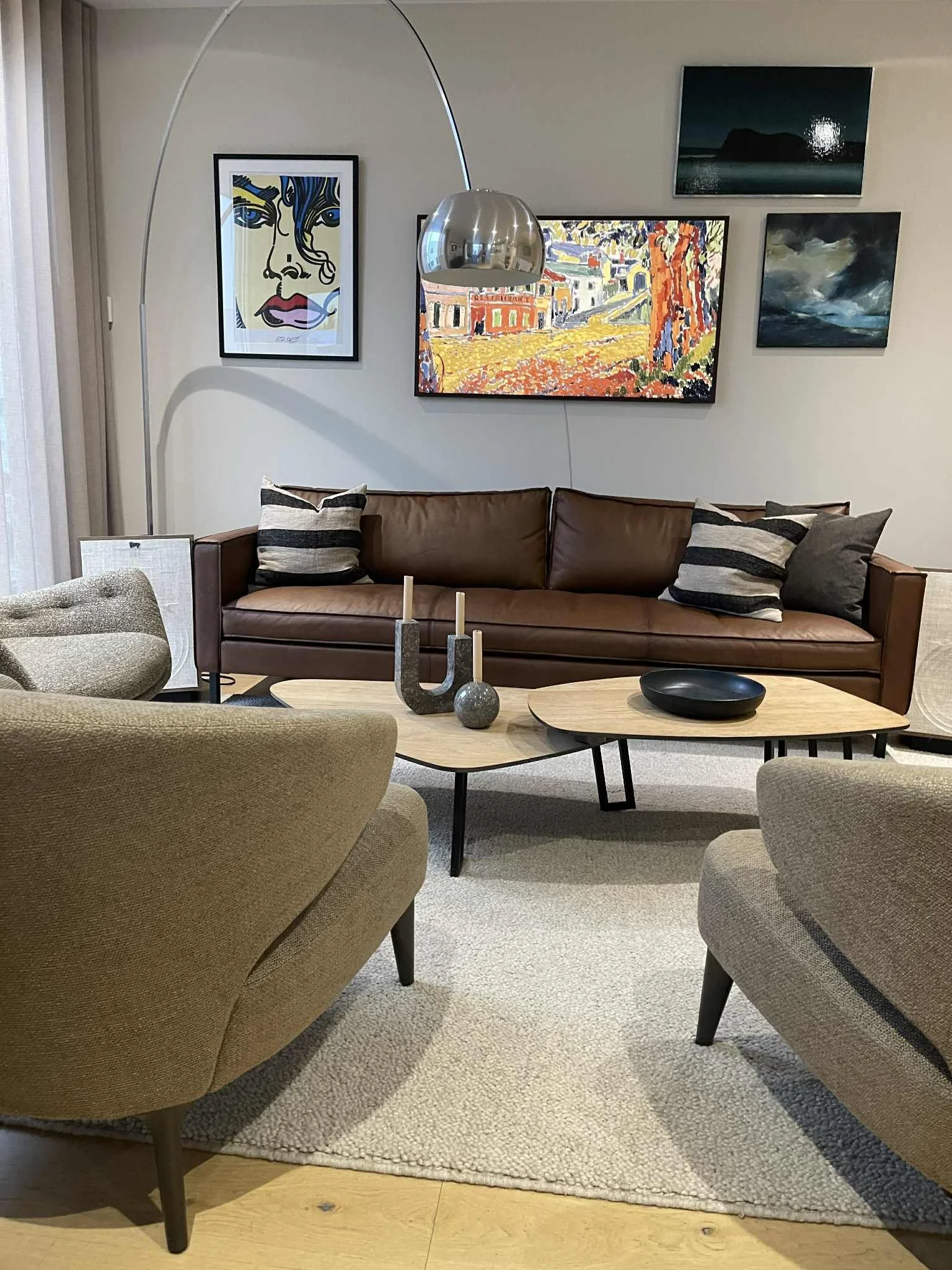 Living room with a brown couch, beige armchair, black coffee table, modern art on the walls, and a silver arc floor lamp.