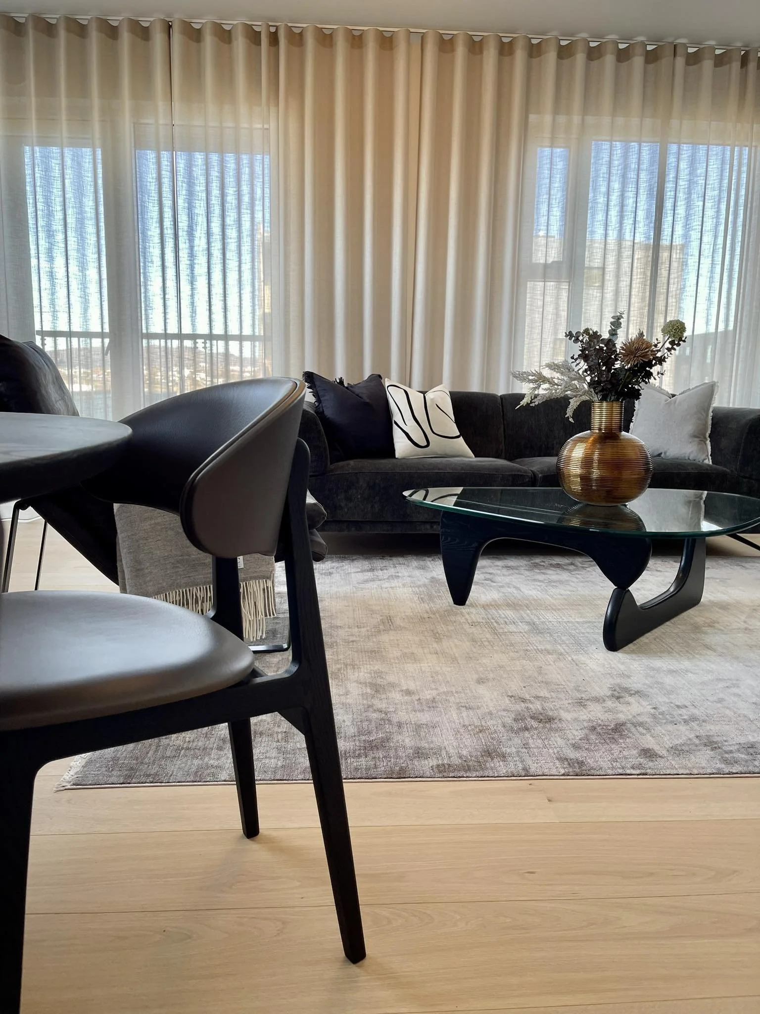 Living room with large sheer curtains, a black sofa with pillows, a glass coffee table with a decorative vase, and dining chairs, with a beige rug on light wood flooring.