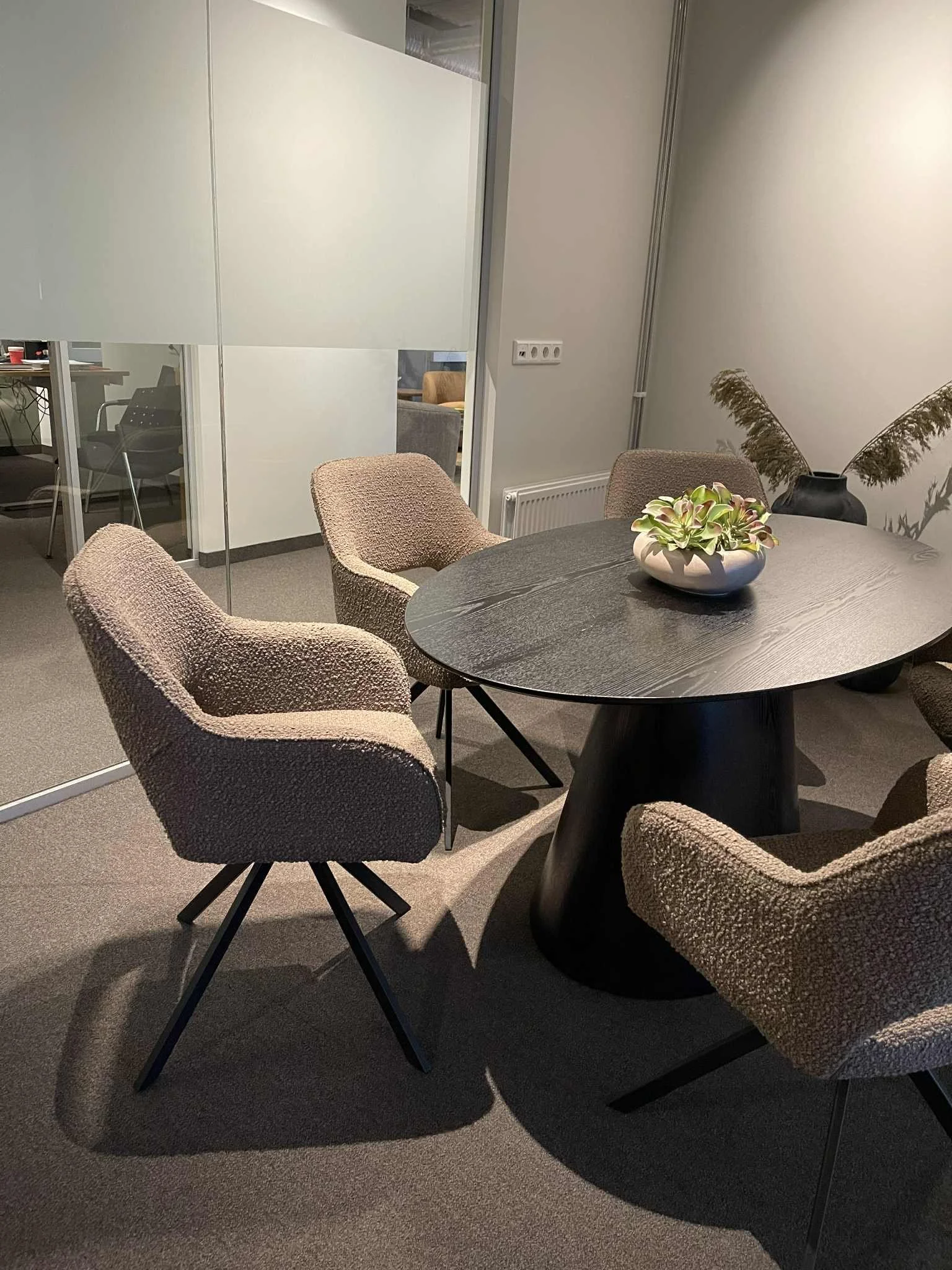 A round black conference table with six beige upholstered chairs, a bowl with green and purple plants, and a black vase with dried brown pampas grass in a modern meeting room.