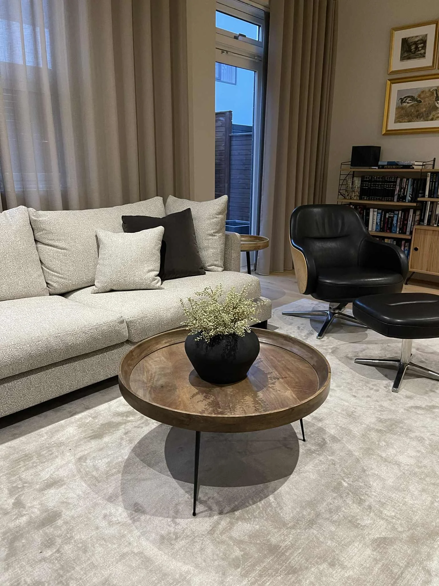 Living room with beige sofa, black and beige cushions, round coffee table with a black vase and white flowers, black lounge chair, wooden bookshelf with books, framed artwork on the wall, window with curtains and a wooden fence outside.