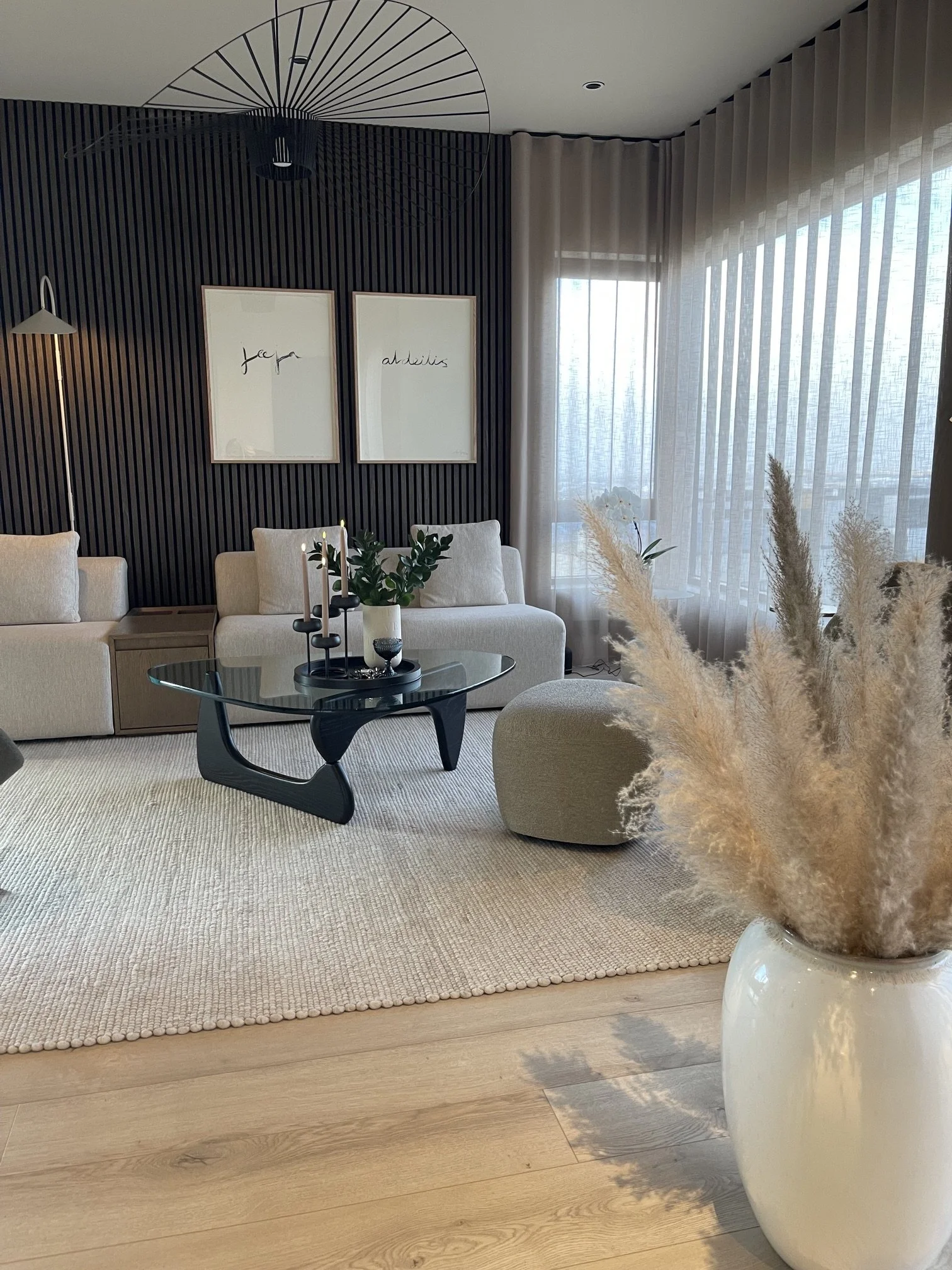 A modern living room with beige sofas, a black coffee table, and a large white vase with pampas grass, featuring a black pop-art inspired ceiling light, black slatted wall with white framed prints, and floor-to-ceiling sheer curtains letting in natur