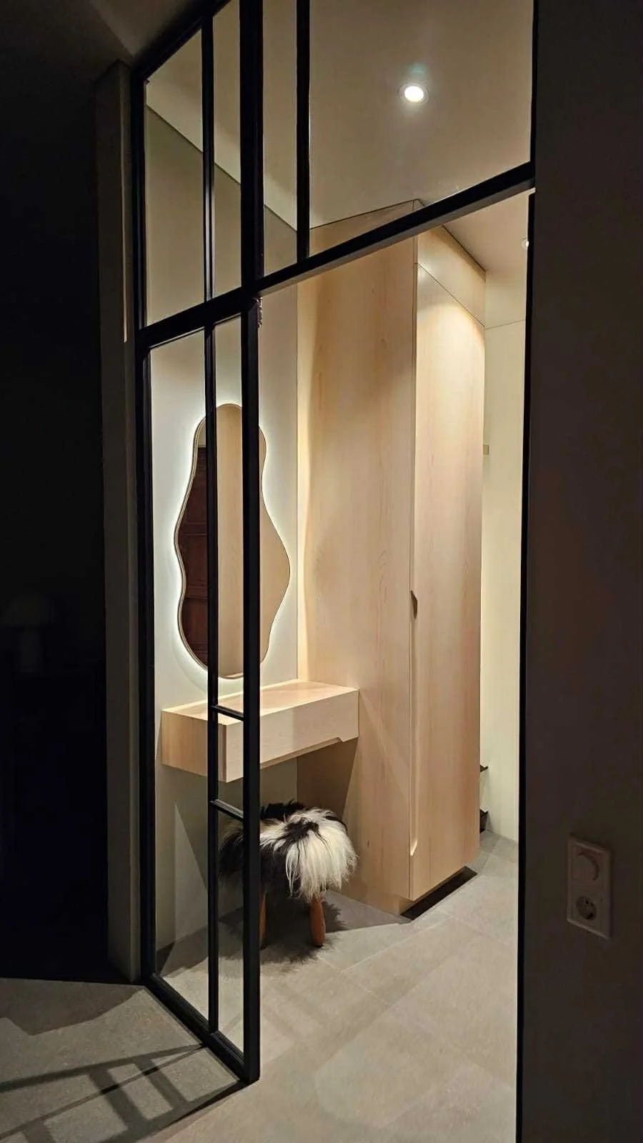 Modern entryway with a light wooden closet, a uniquely shaped mirror, a small floating desk, and a patterned dog with fluffy tail standing on the floor.