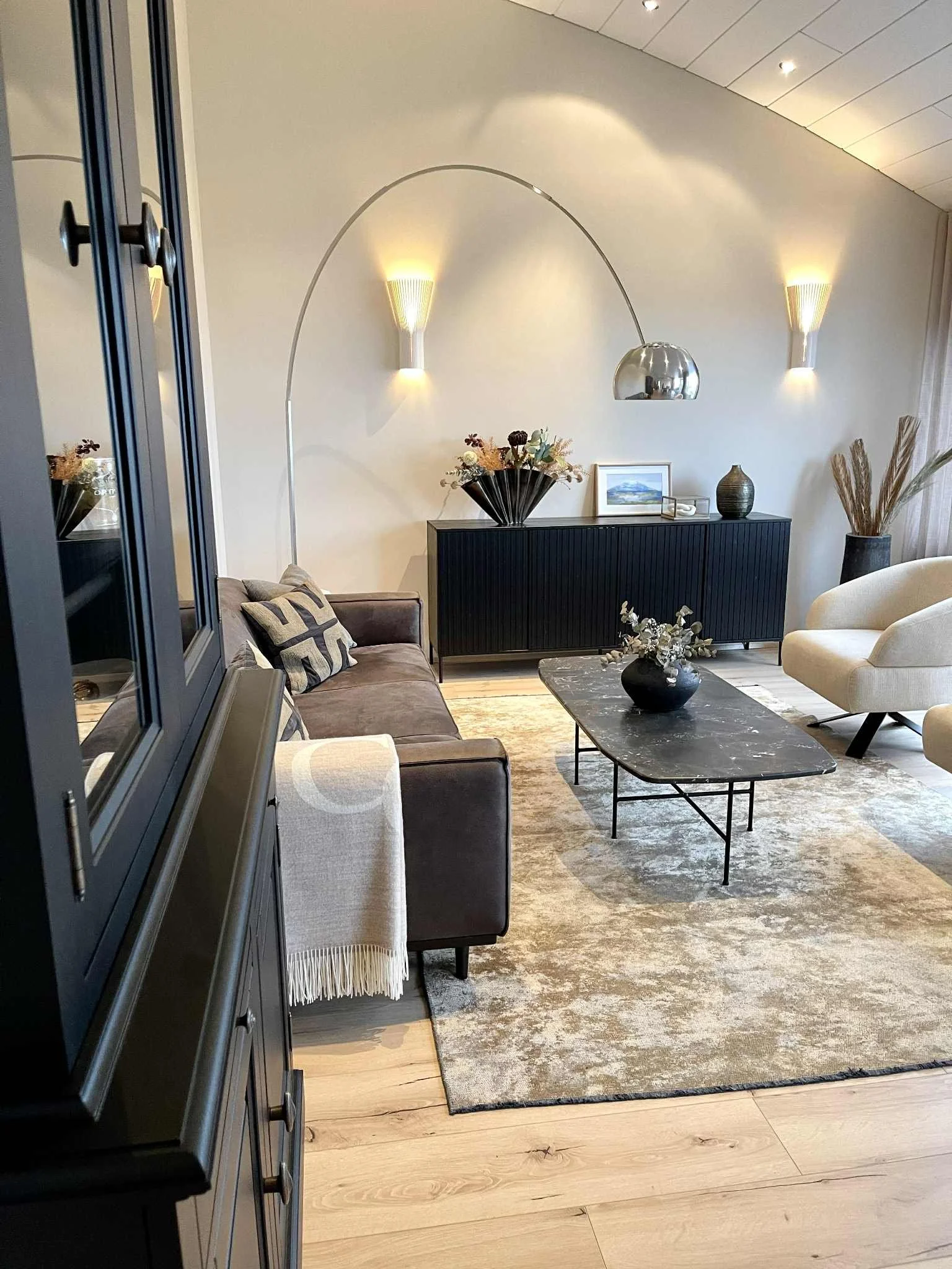 Living room with cream-colored walls and a sloped ceiling, featuring modern lighting, a black sideboard with a floral arrangement, a framed picture, vases, and a sculpture, with beige and dark furniture, a marble coffee table, and a large rug.