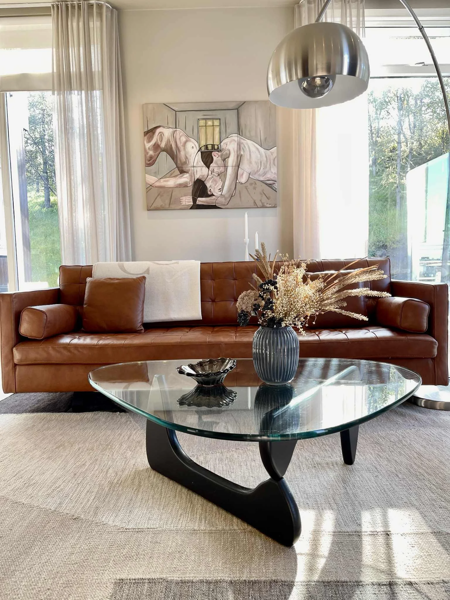 Living room with a brown leather sofa, a glass coffee table, a decorative vase with dried flowers, and a wall painting of two nude women bowing their heads, with large windows and sheer curtains letting in natural light.