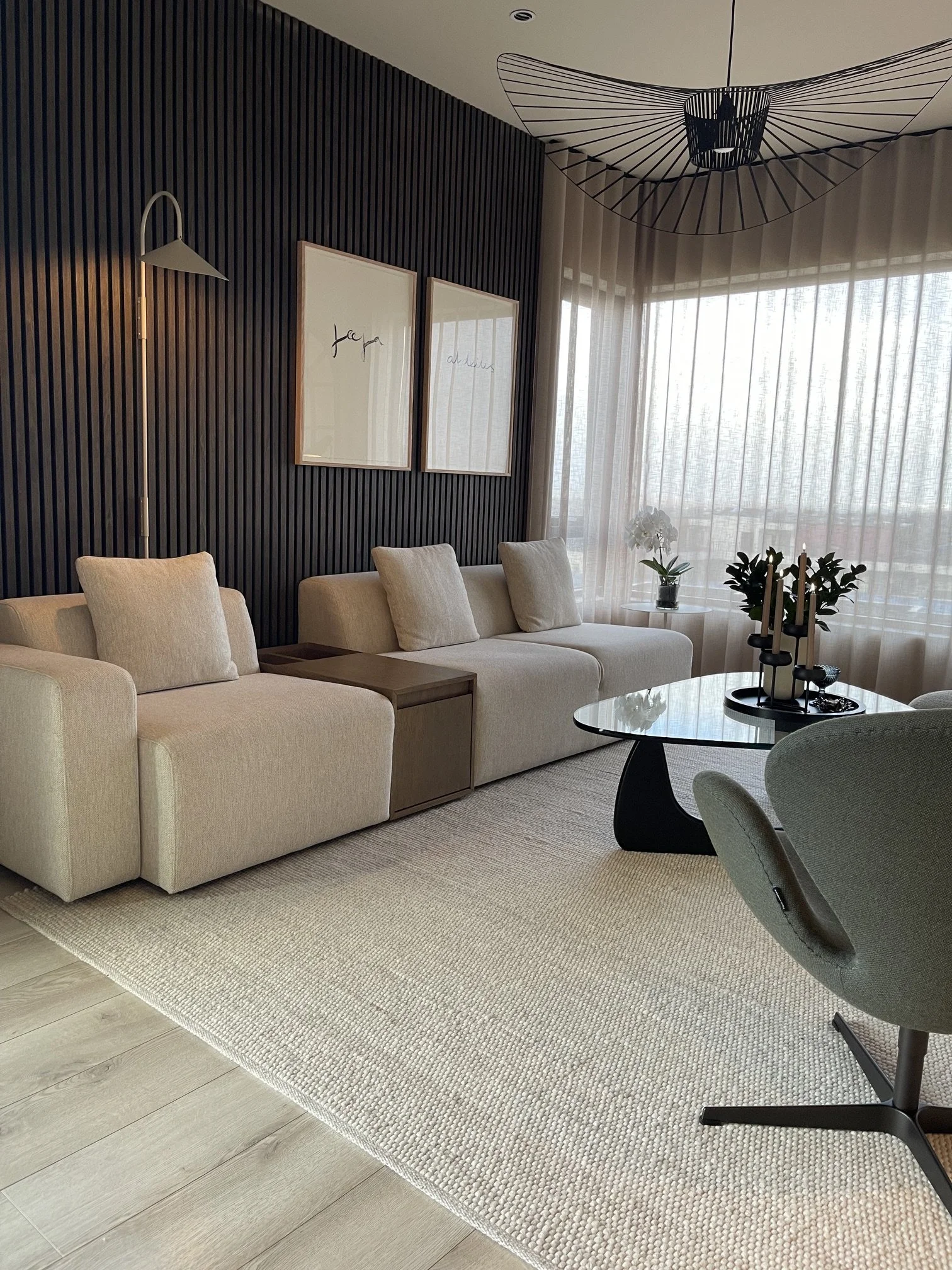 Living room with beige sofa, a round black coffee table, window with sheer curtains, and wall decorations.