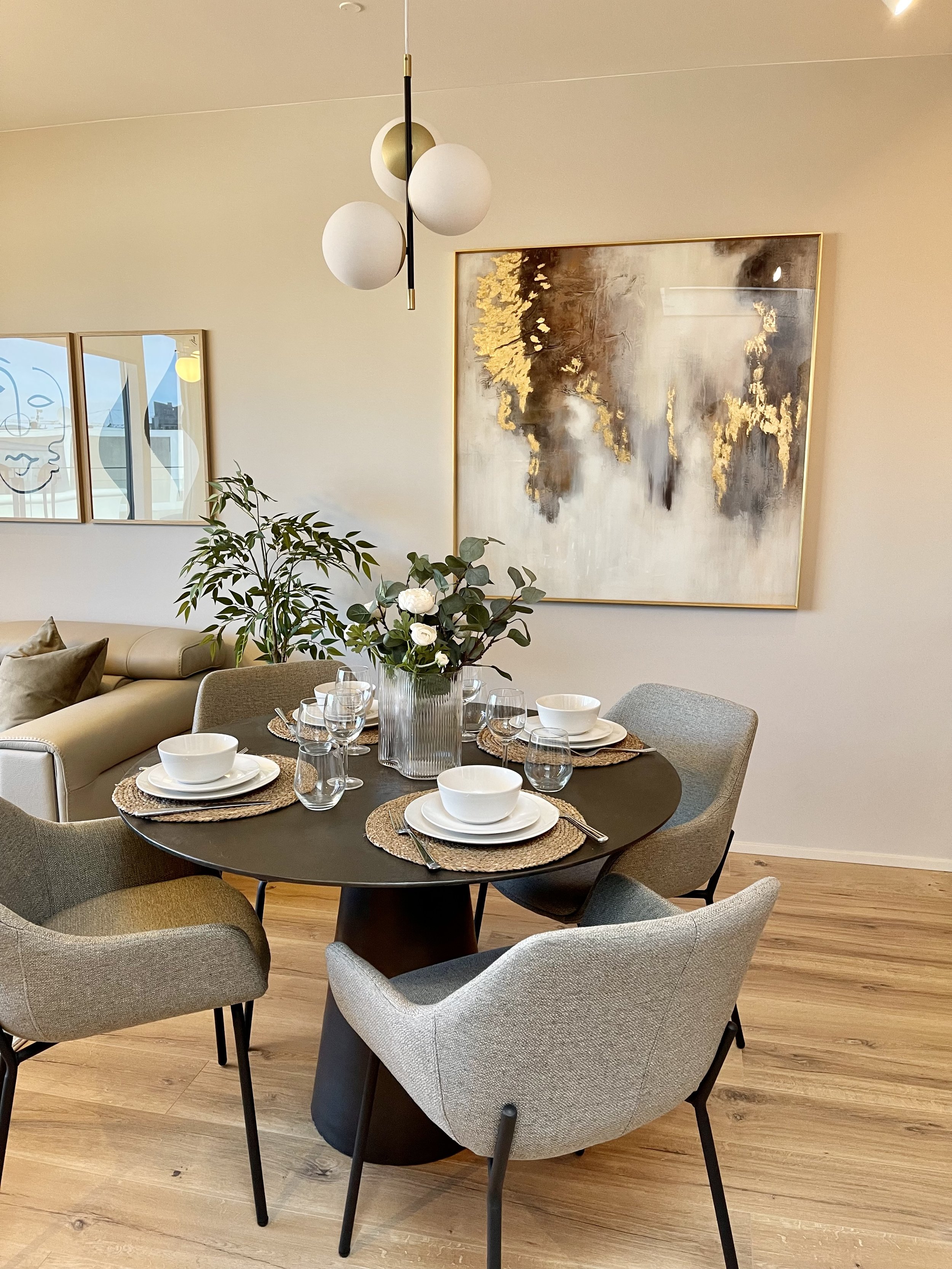 A dining room with a round black table set with white bowls, plates, and glassware, surrounded by six gray upholstered chairs, with a large abstract painting on the wall and a glass vase with greenery and white flowers in the center of the table.
