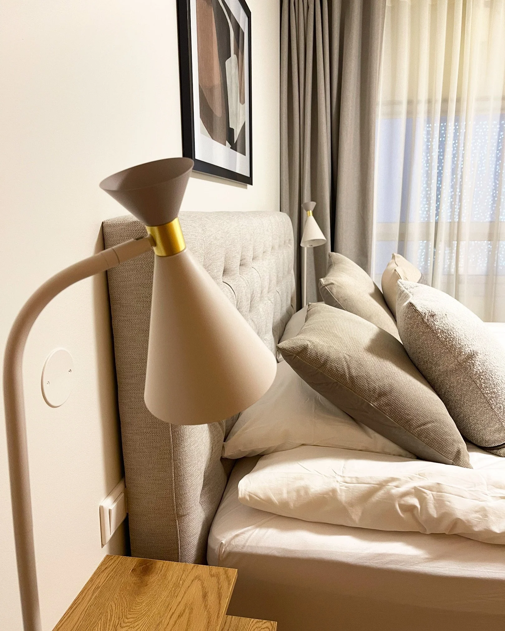 A hotel room with a beige headboard, multiple pillows, a bed with white linen, a bedside lamp, a window with beige curtains, and abstract art on the wall.