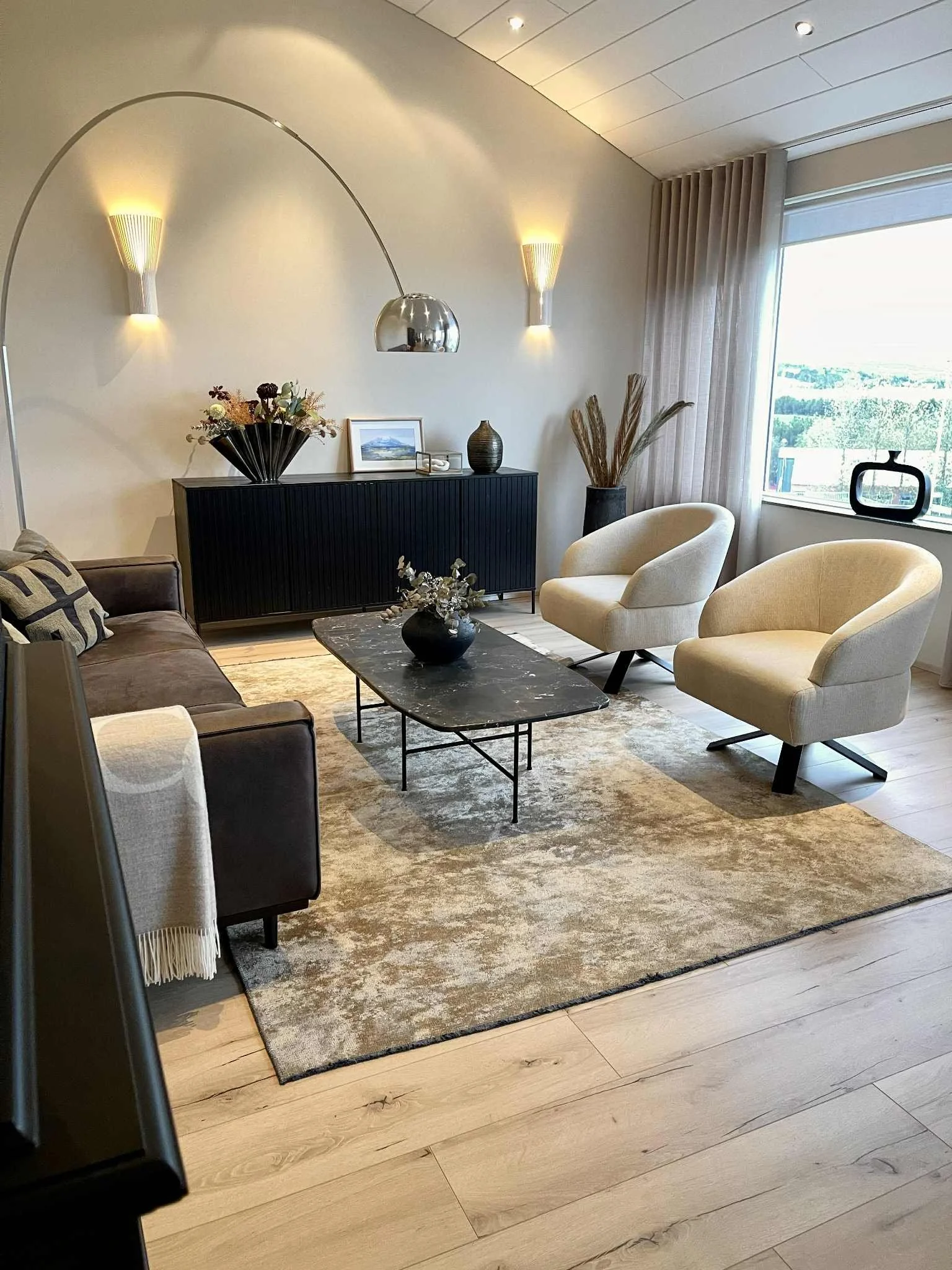 A living room with a dark sofa, two light-colored armchairs, a marble coffee table, a black sideboard with decorative items, a vase with dried plants, a large window with sheer curtains, and wall-mounted lights.
