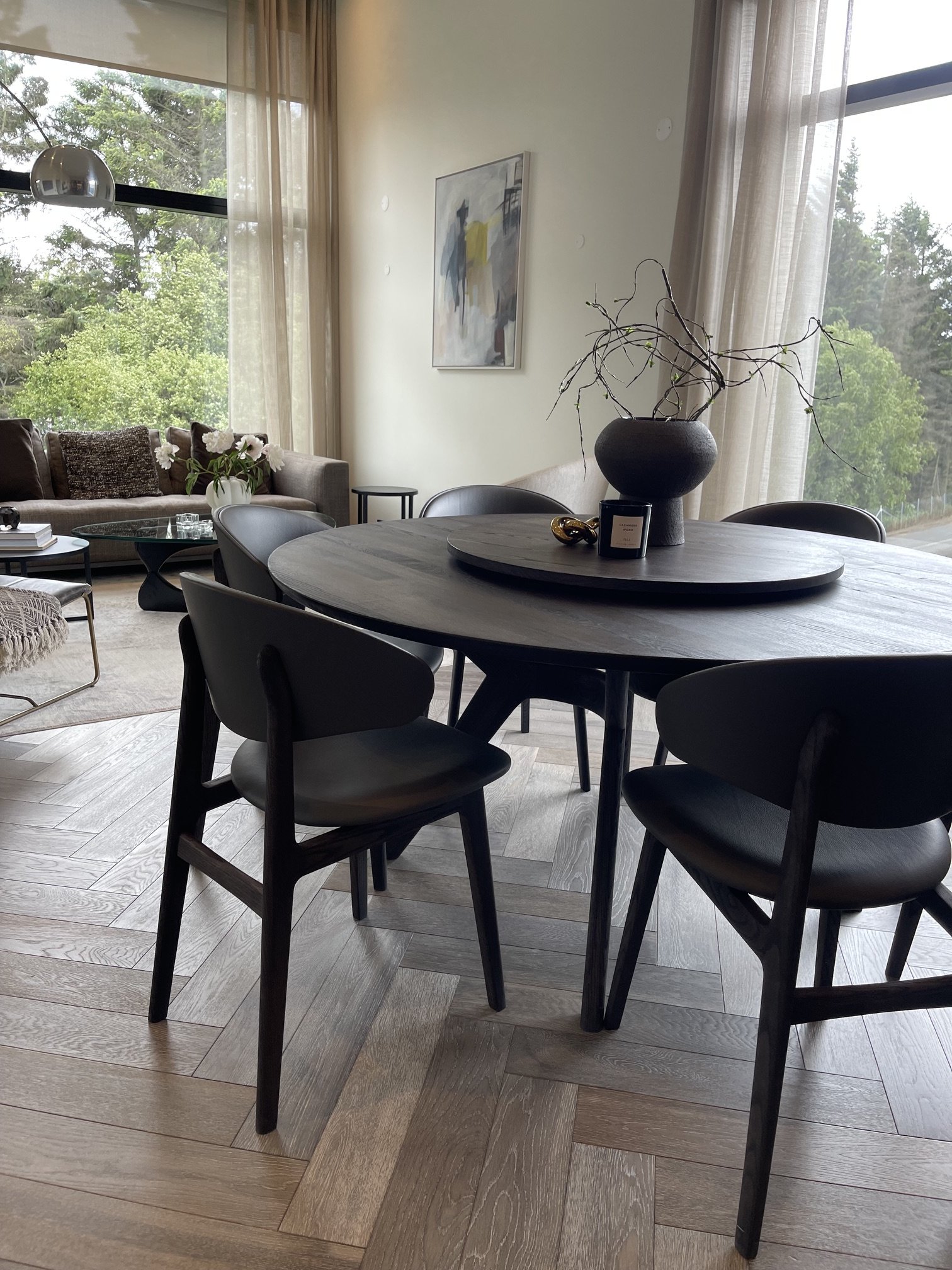 A modern living room with large windows, a round dining table with black chairs, a beige sofa with pillows, a glass coffee table, and decorative vases with flowers, with outdoor greenery visible through the windows.