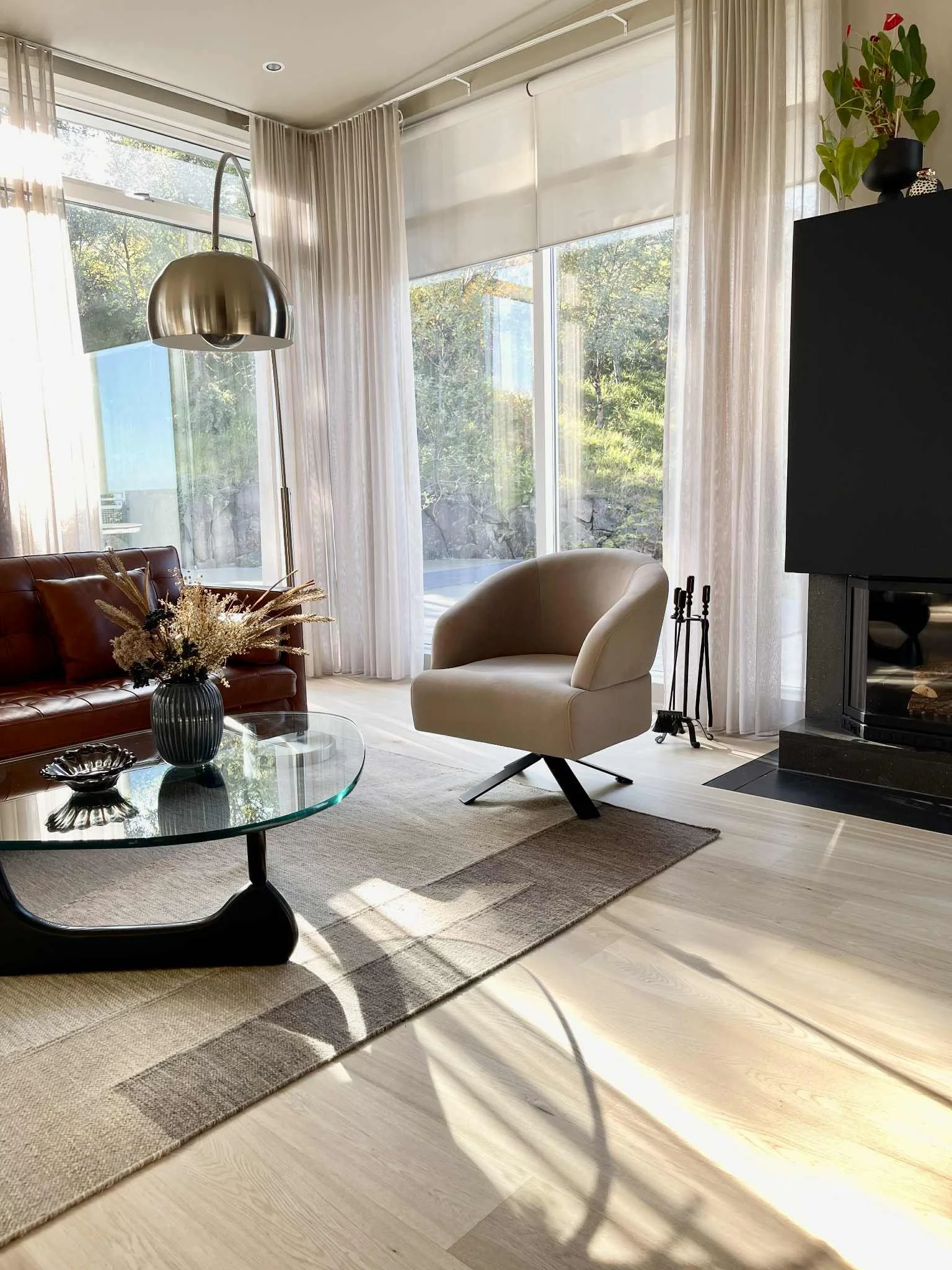 Bright living room with large windows, white curtains, beige armchair, red leather sofa, glass coffee table with decorative items, grey area rug, and a modern fireplace, with sunlight streaming in.