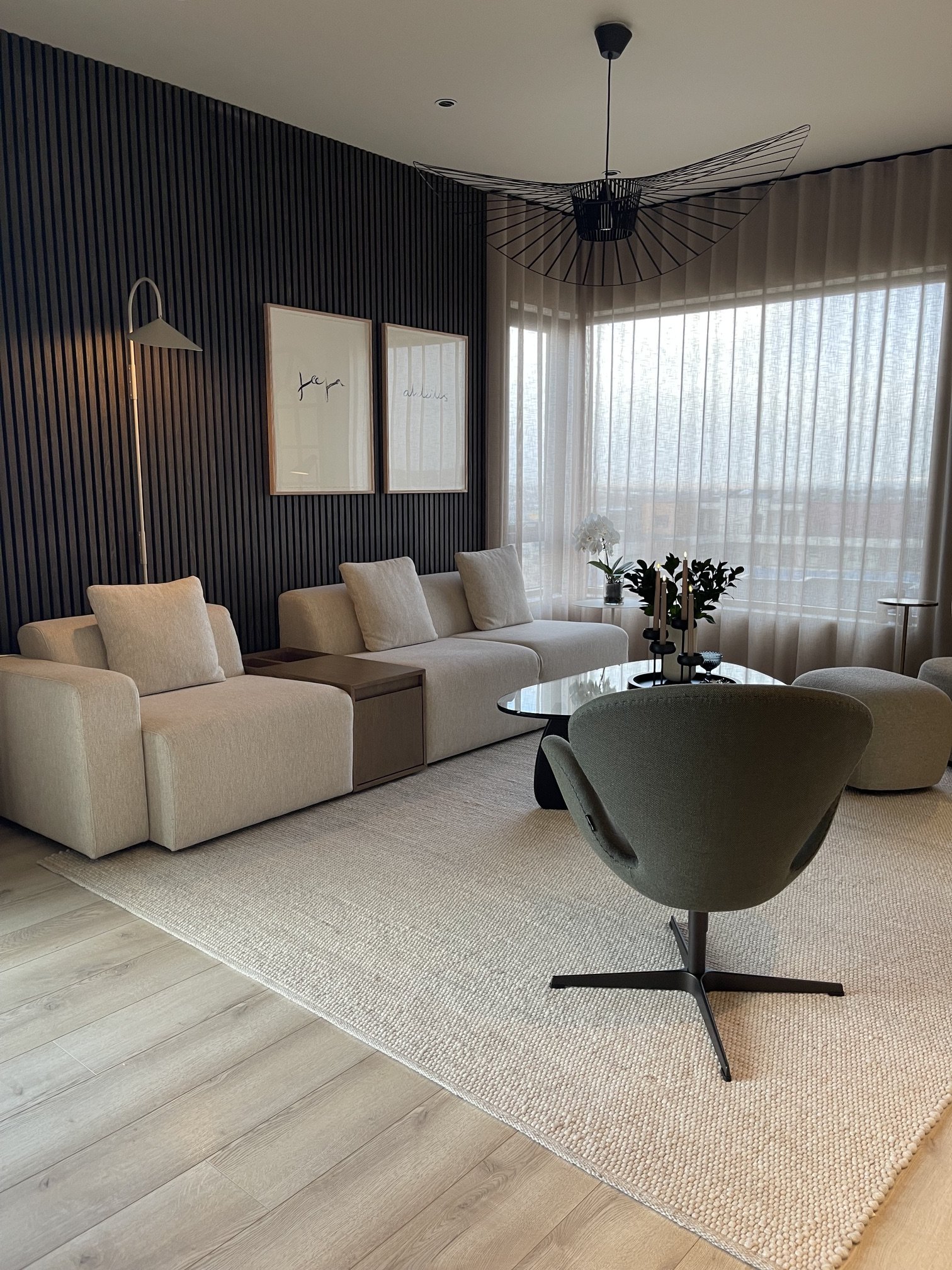 Modern living room with beige sofa, black wireframe pendant light, white curtains, potted orchid and greenery, coffee table with candles, wall with two framed artworks, black wall panel, standing lamp, and light wood flooring.