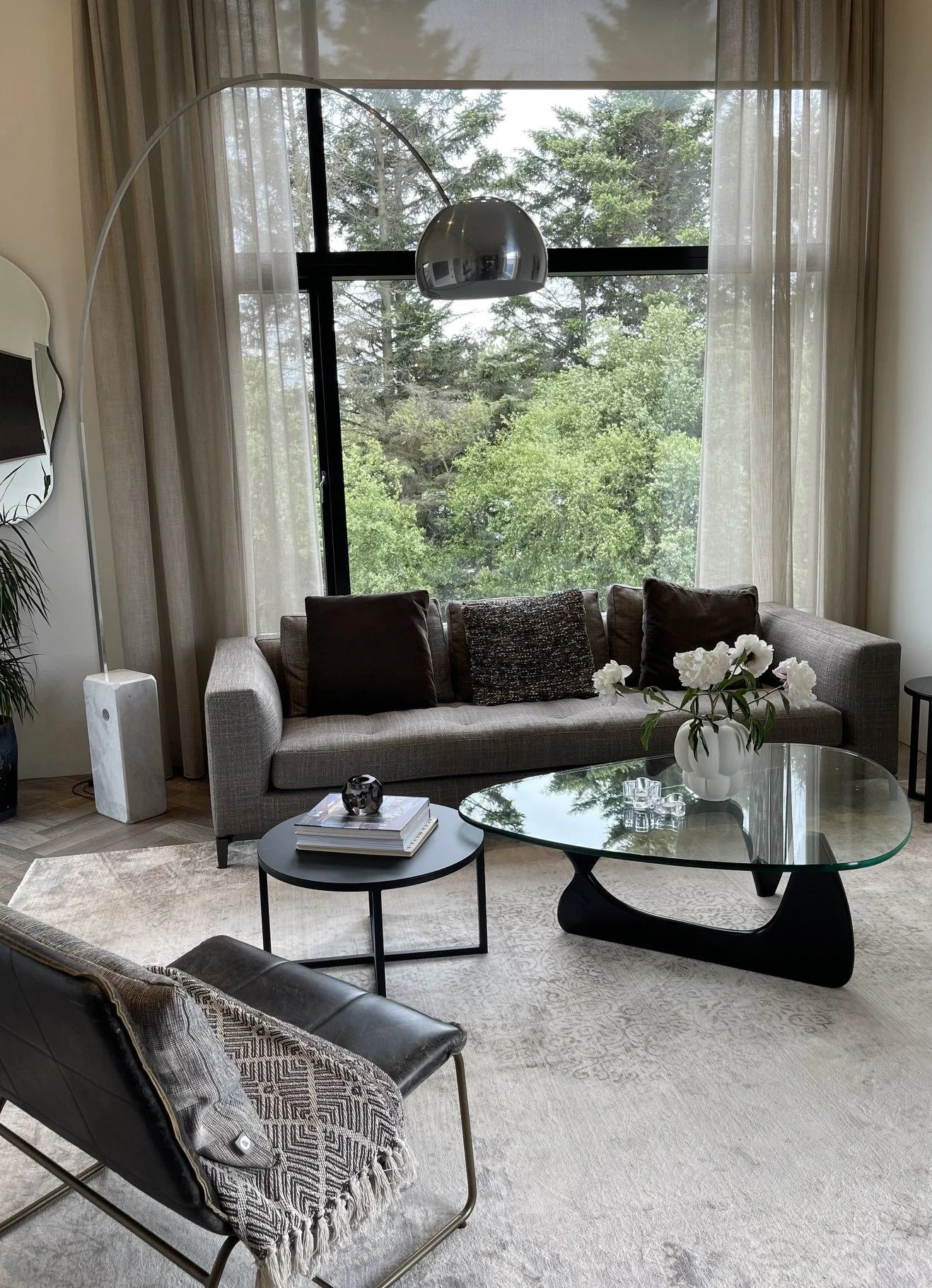 Living room with beige sofa, dark throw pillows, glass coffee table with white flowers, black side tables, modern armchair with patterned throw blanket, large window with curtains showing trees outside, decorative mirror, and a metallic floor lamp.