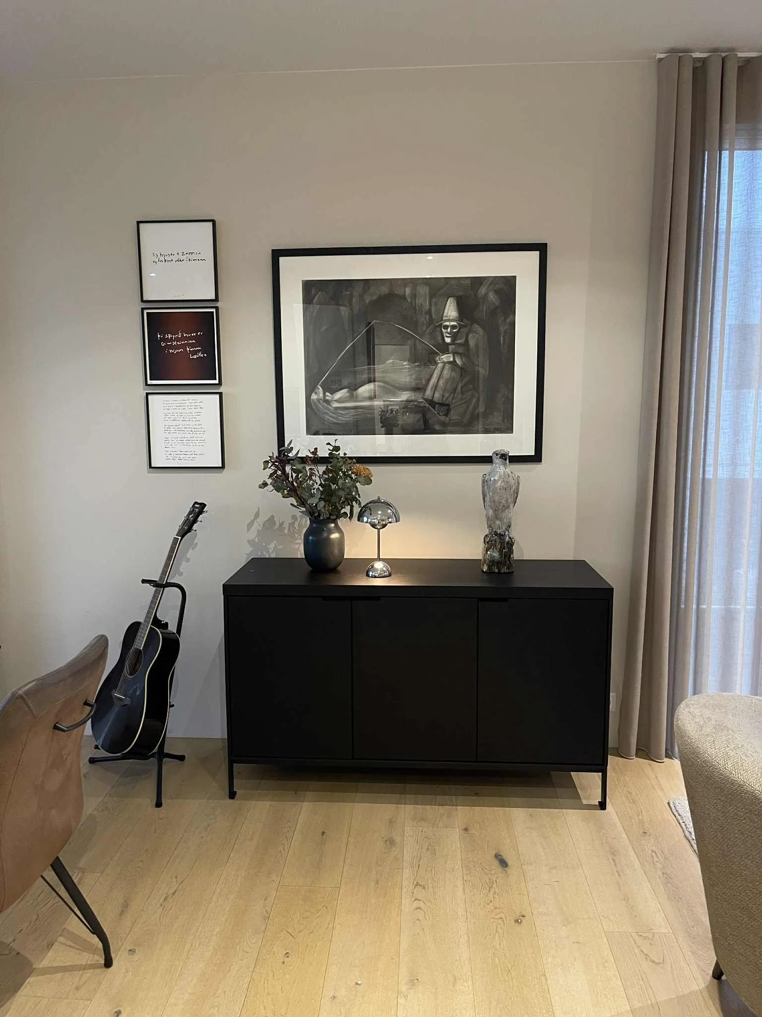 A black sideboard with decorative items on top, including a plant in a dark vase, a small shiny lamp, and a tall, textured glass vase. A black and white framed artwork hangs above. To the left, a guitar leans against a chair. To the right, curtains a
