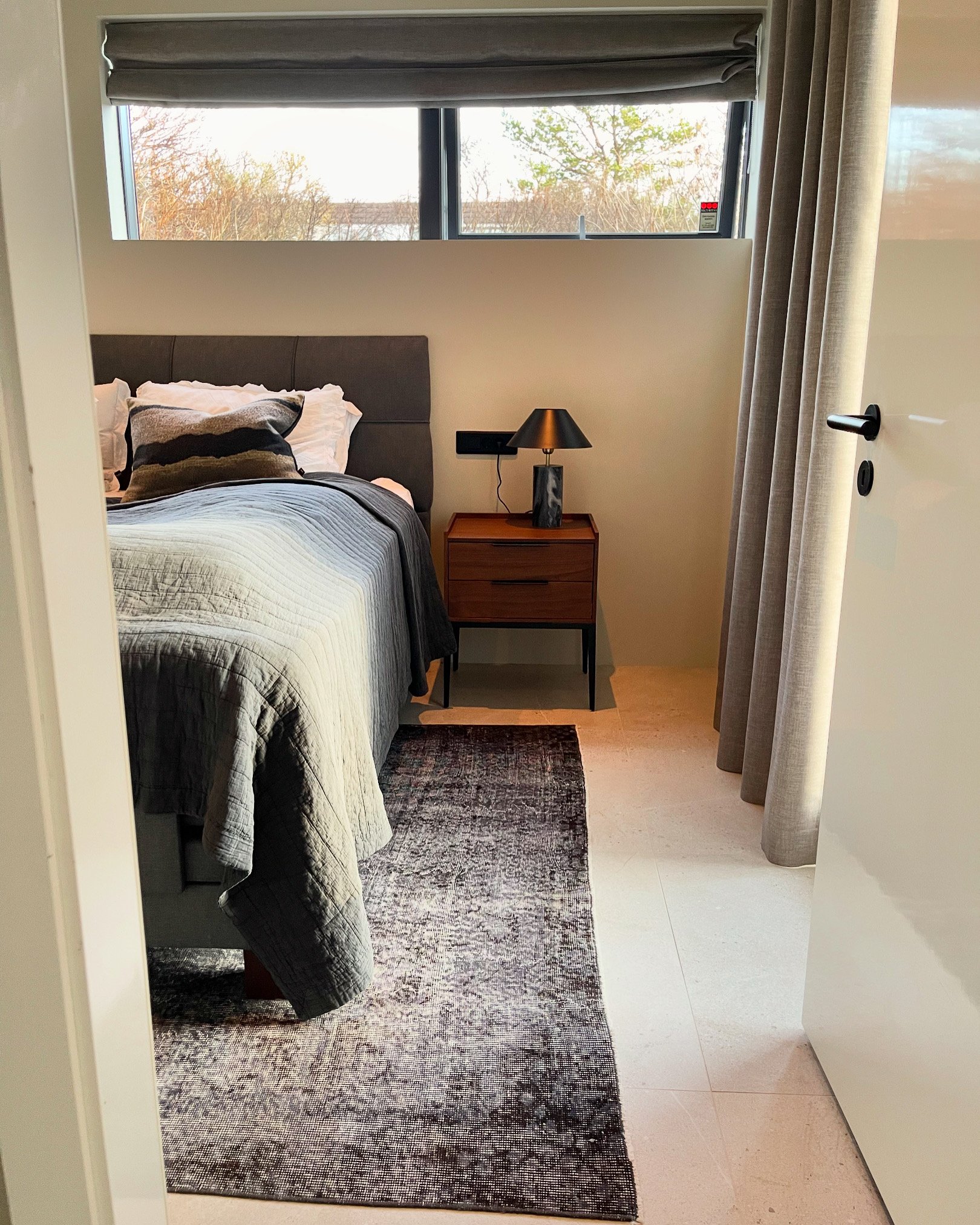 A bedroom viewed through a doorway, featuring a bed with a dark headboard, white bedding, and decorative pillows. A small wooden night table with a black and gold lamp is beside the bed. There is a window above the bed with an outside view of trees a