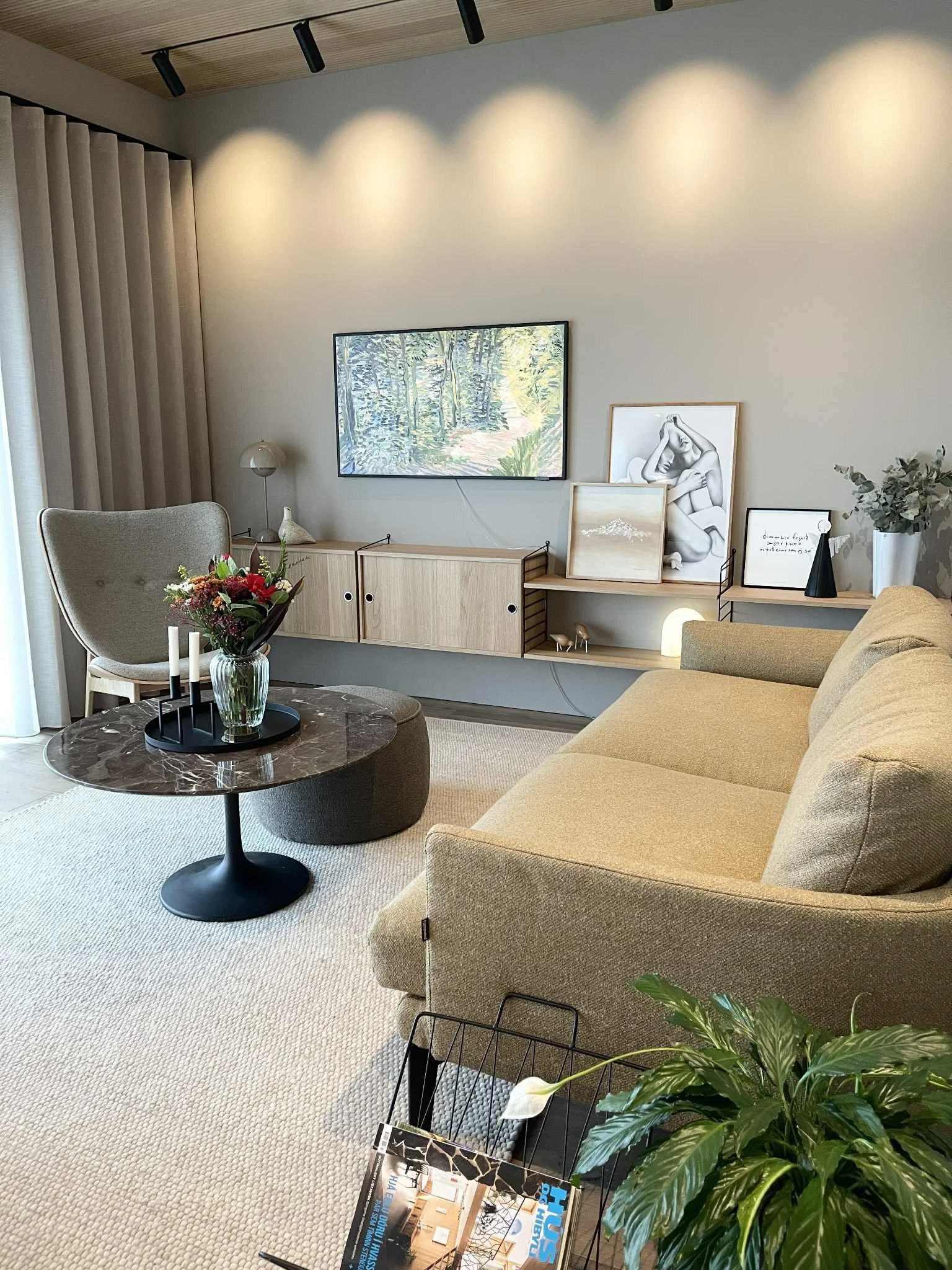 A cozy living room with a beige sofa, a black marble coffee table with a vase of flowers, a light-colored armchair, a wooden TV stand with framed artwork, and a flat-screen TV on the wall. There is also a plant, a magazine rack, a lamp, and a wall wi