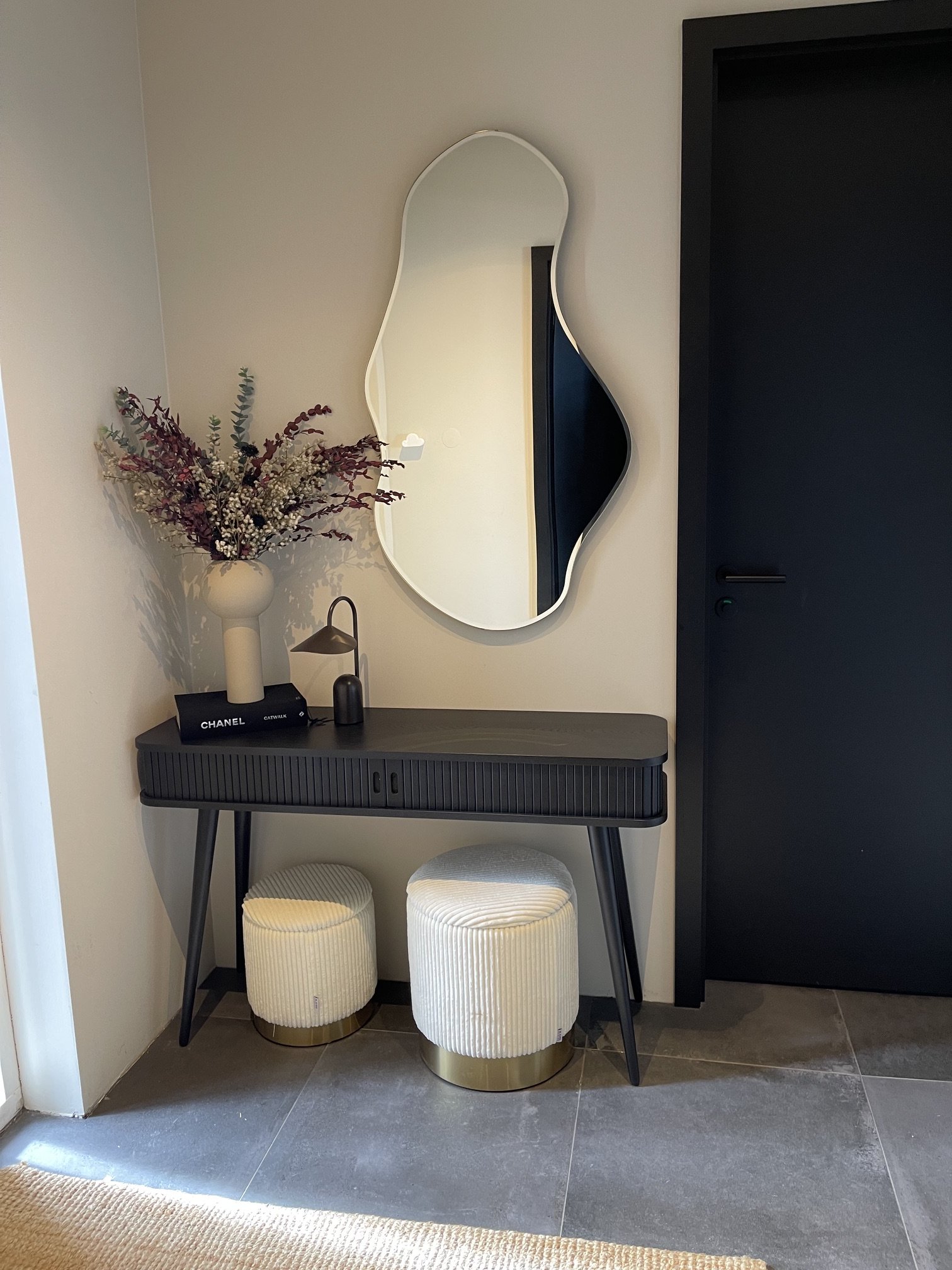 A modern console table with a black ribbed surface and tapered black legs. On top, there is a beige vase with a bouquet of dry flowers, a small black box labeled 'CHANEL CATWALK,' a black lamp, and a black bottle. There are two white, ribbed, cylindr