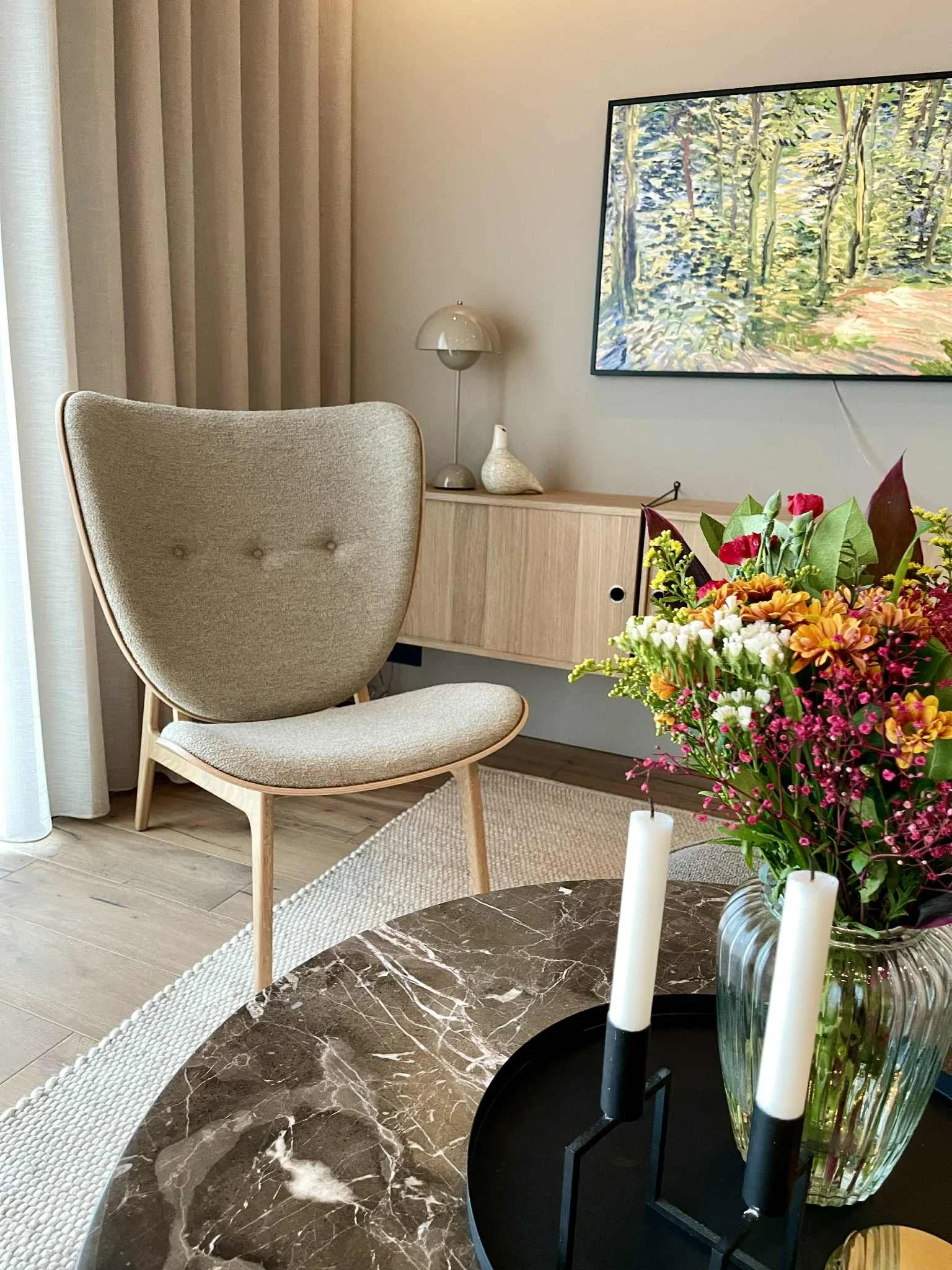 A modern living room corner with a beige upholstered armchair, a wooden sideboard with a small lamp and decorative item, a large framed landscape painting on the wall, a bouquet of colorful flowers on a black tray, and candles on a marble coffee tabl