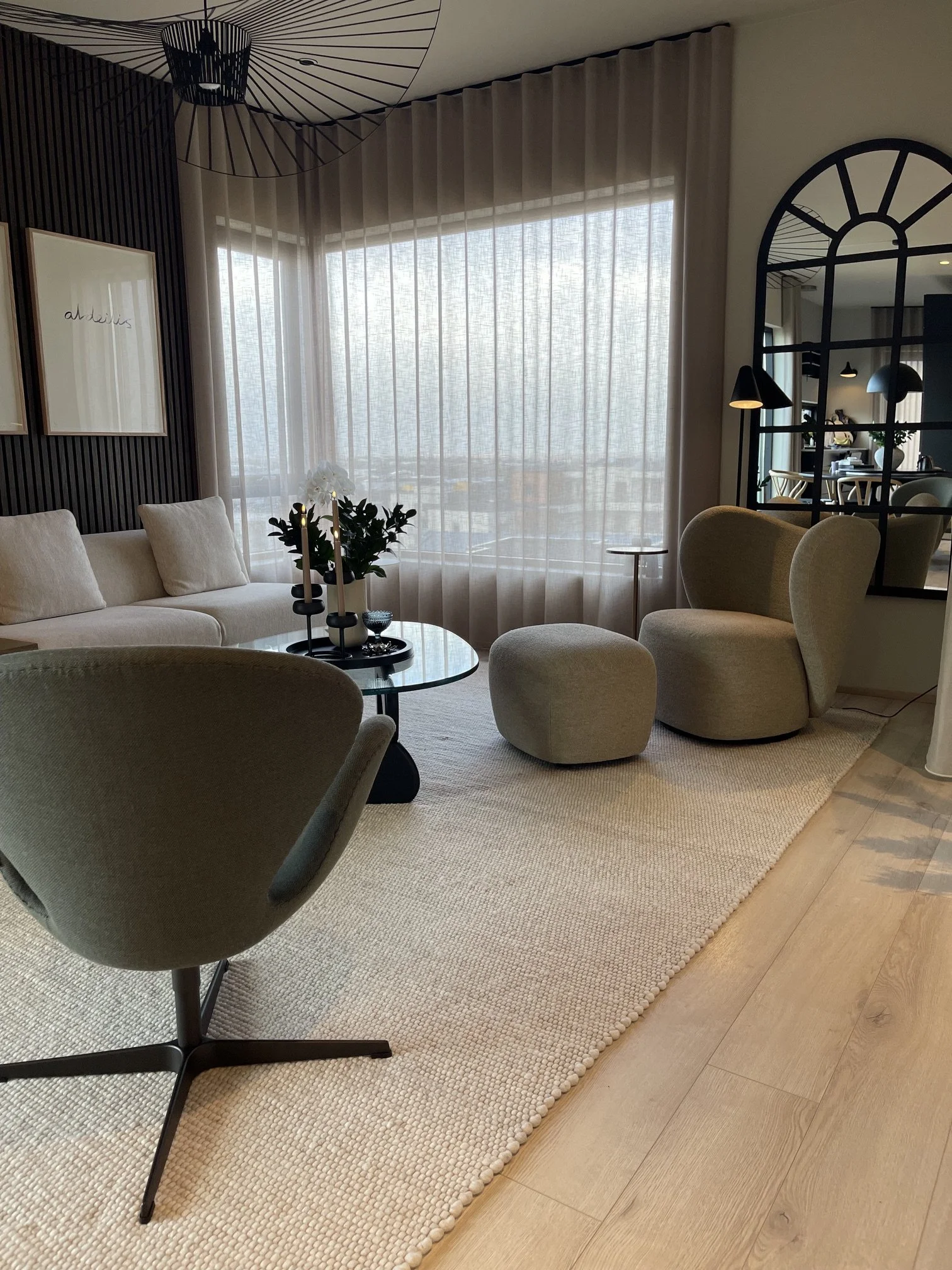 Modern living room with beige upholstered furniture, a glass coffee table with black candles and white orchids, a large sheer window with beige curtains, a textured black accent wall with framed artwork, and a patterned beige rug with wooden flooring