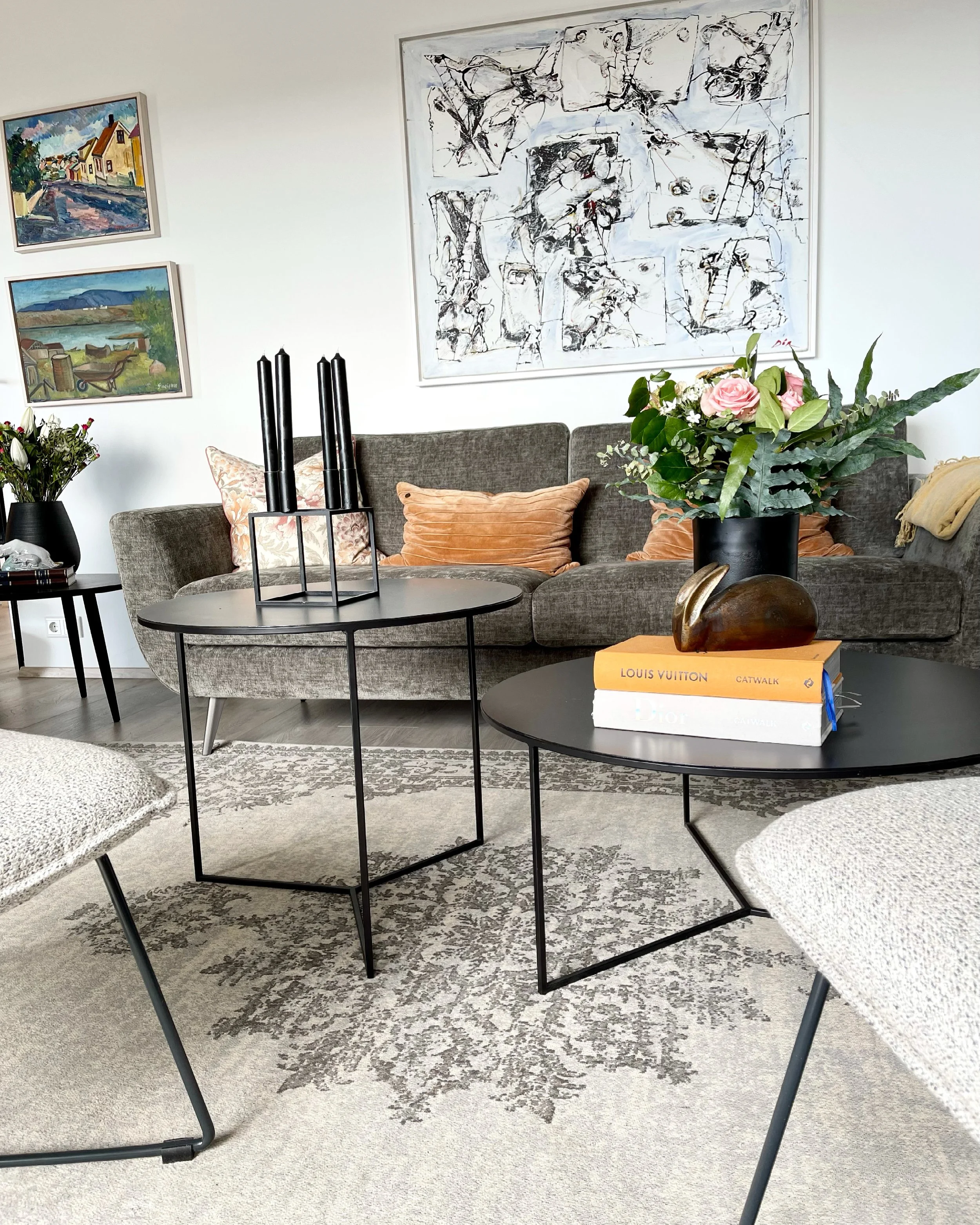 A living room with a gray sofa adorned with orange and patterned pillows, a black coffee table with a large plant in a black pot, and a side table with a smaller plant. The wall features three paintings: two landscape scenes and one abstract piece. A