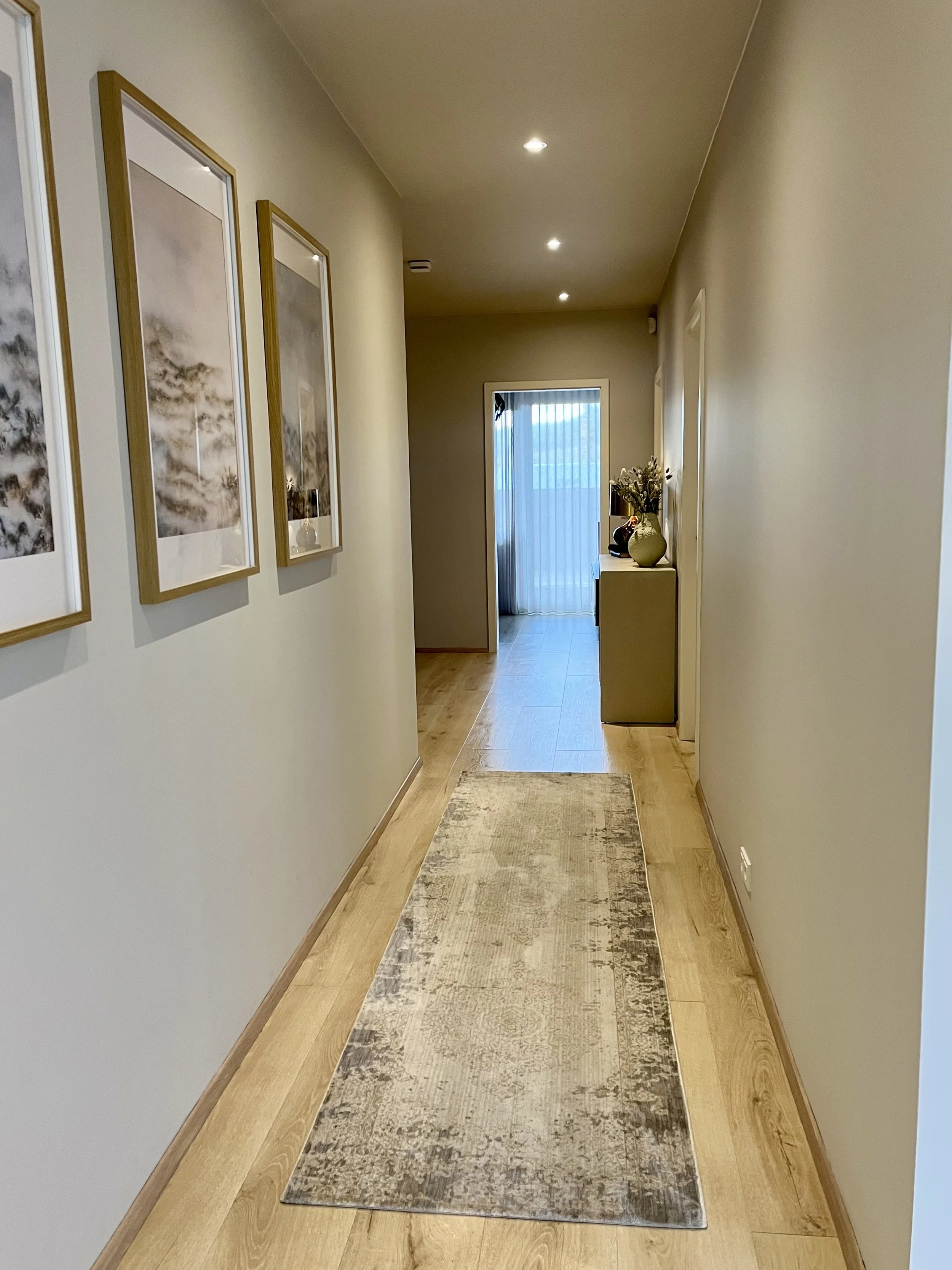A hallway with wooden flooring, beige walls, framed artwork on the left, a runner rug, and a side table with vases and flowers near a sliding glass door with curtains.
