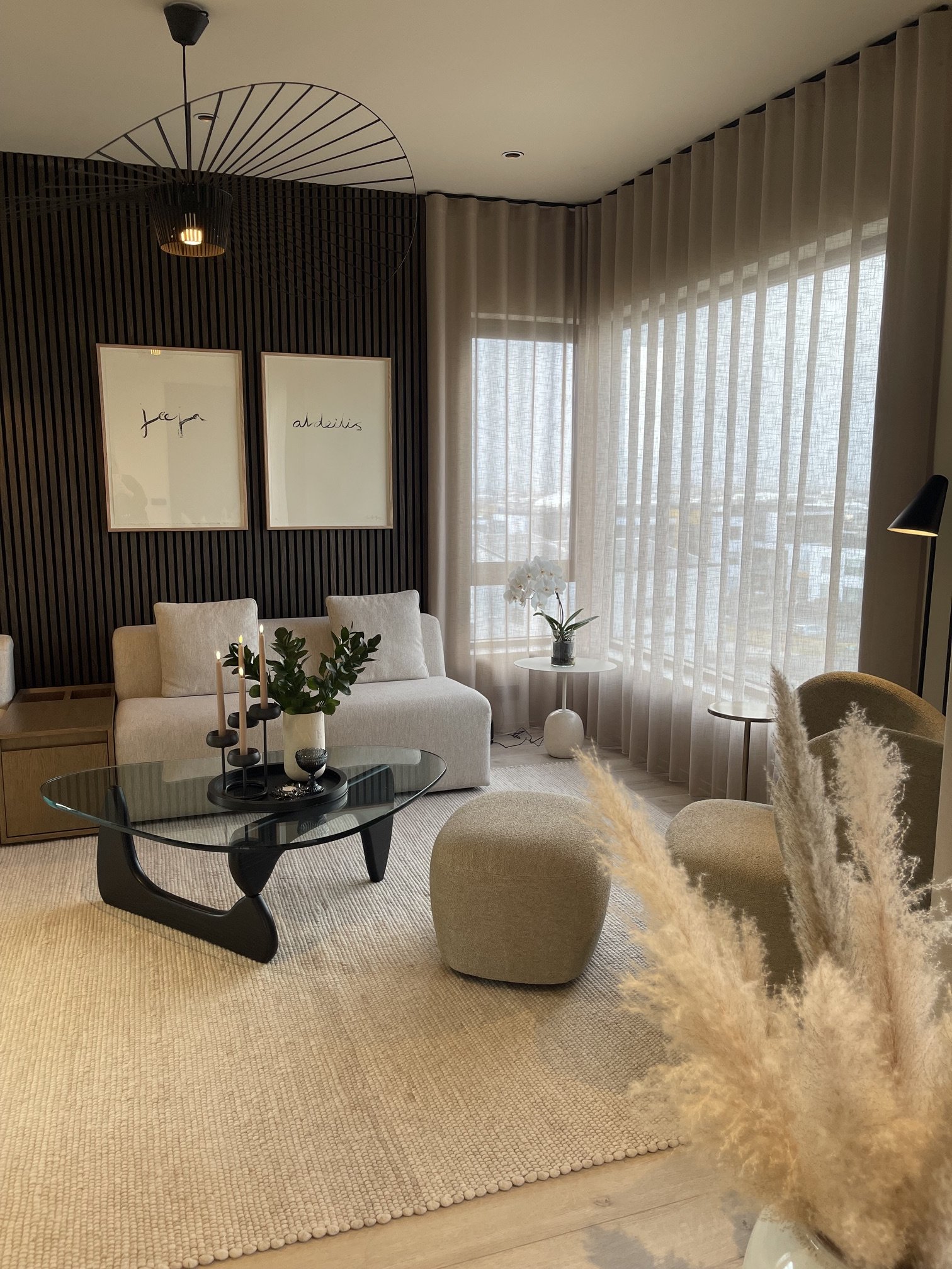 A cozy living room with a beige carpet, a white sofa, a black glass coffee table with candles and a green plant, beige poufs, light-colored curtains, framed wall art, a floor lamp, and a potted orchid near the window, with a modern black and gold cha