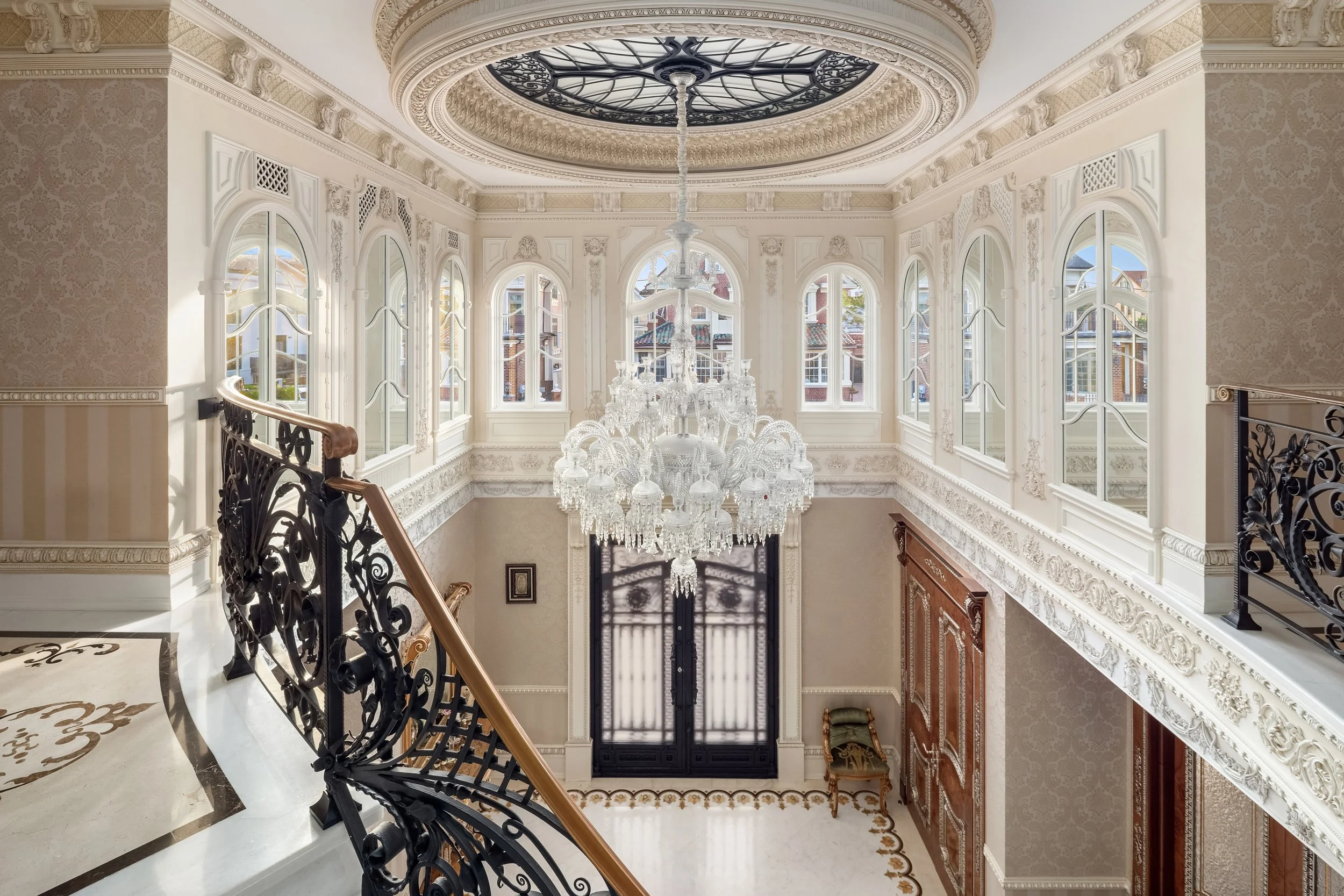Interior view of a luxurious staircase hall with a chandelier, ornate white mouldings, large arched windows, and a decorative ceiling with a glass and wrought iron design.