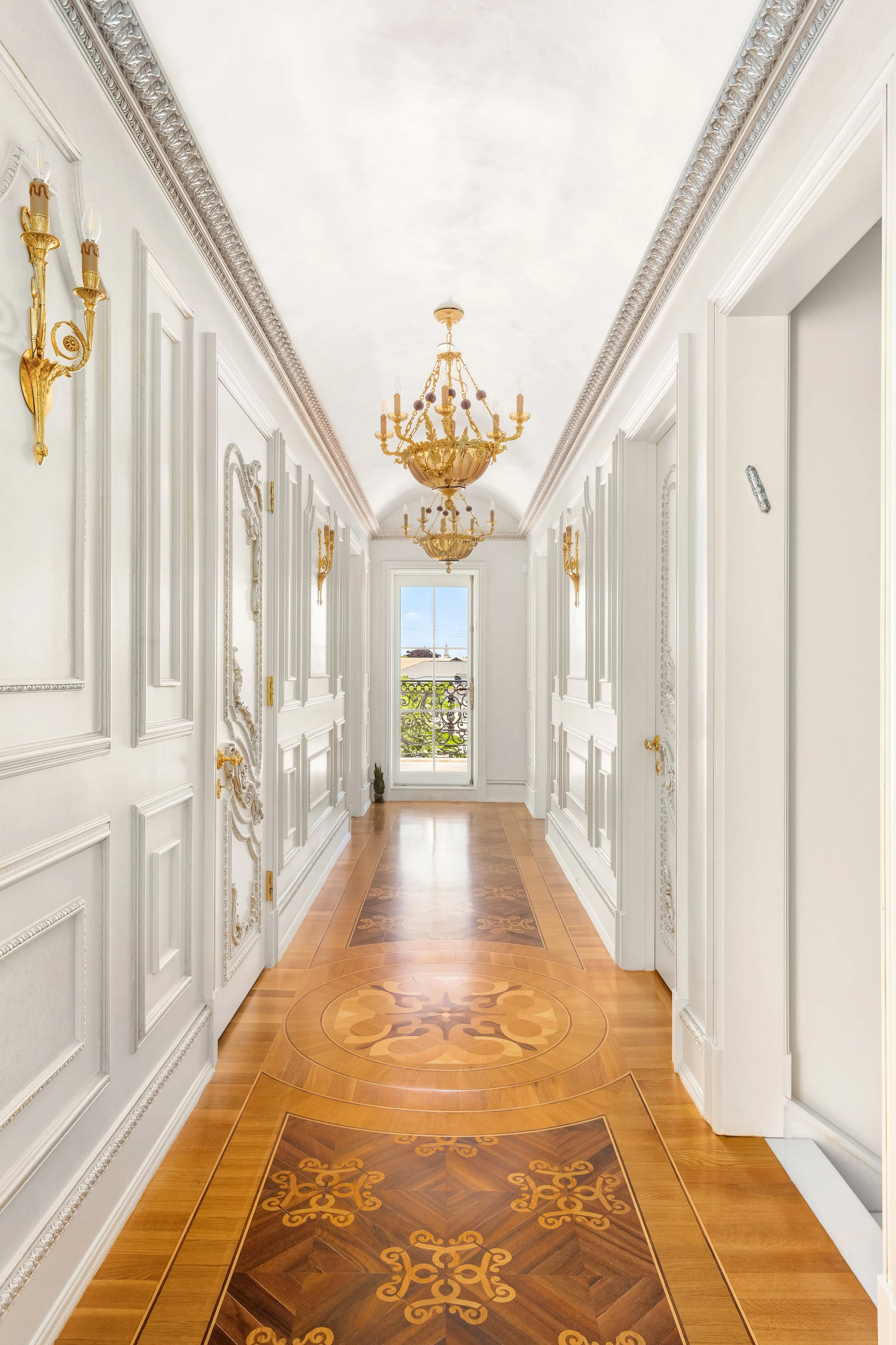 A luxurious hallway with intricate white wall paneling, gold accents, and a wooden floor with ornate inlay patterns. It features two glass chandeliers hanging from a white ceiling, and there's a window at the end of the hall with a balcony outside.