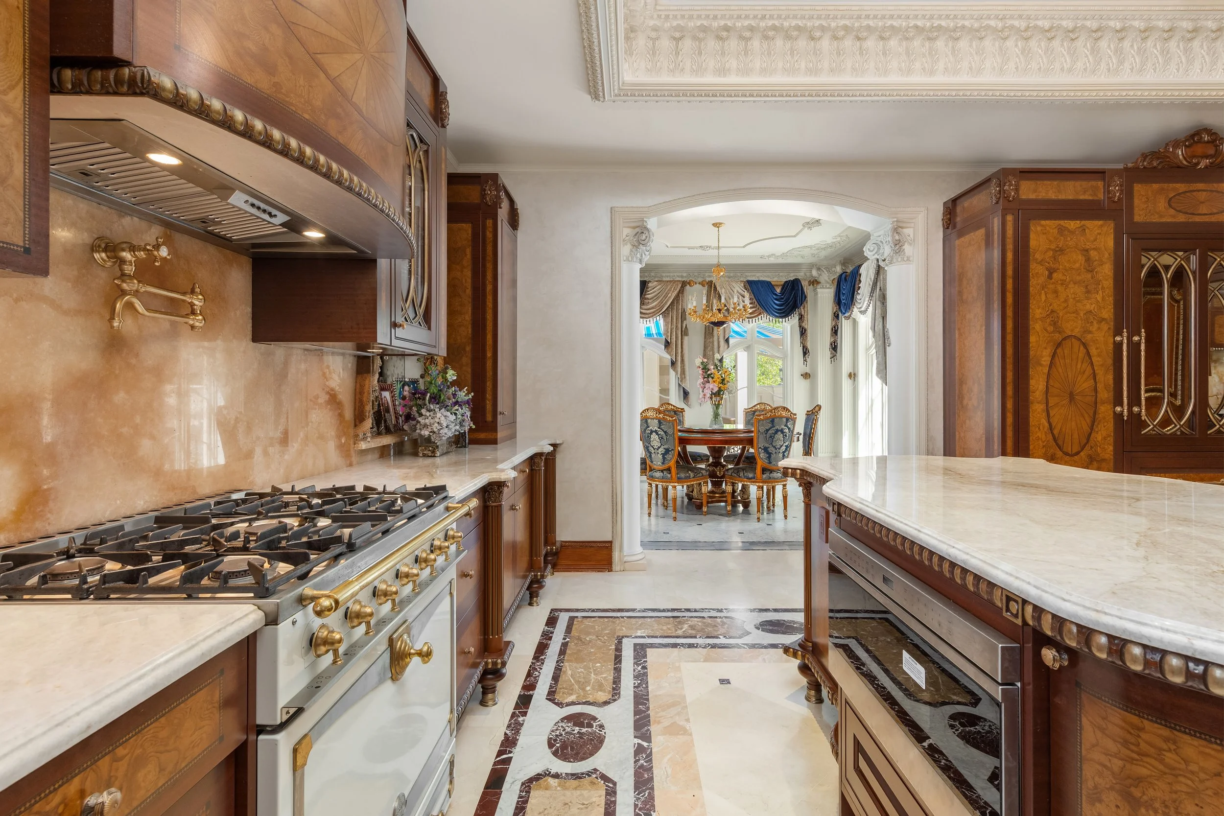 A luxurious kitchen with marble countertops, wooden cabinets, a gas stove, and a dining area with a round table and ornate chairs, brightened by large windows with drapes.
