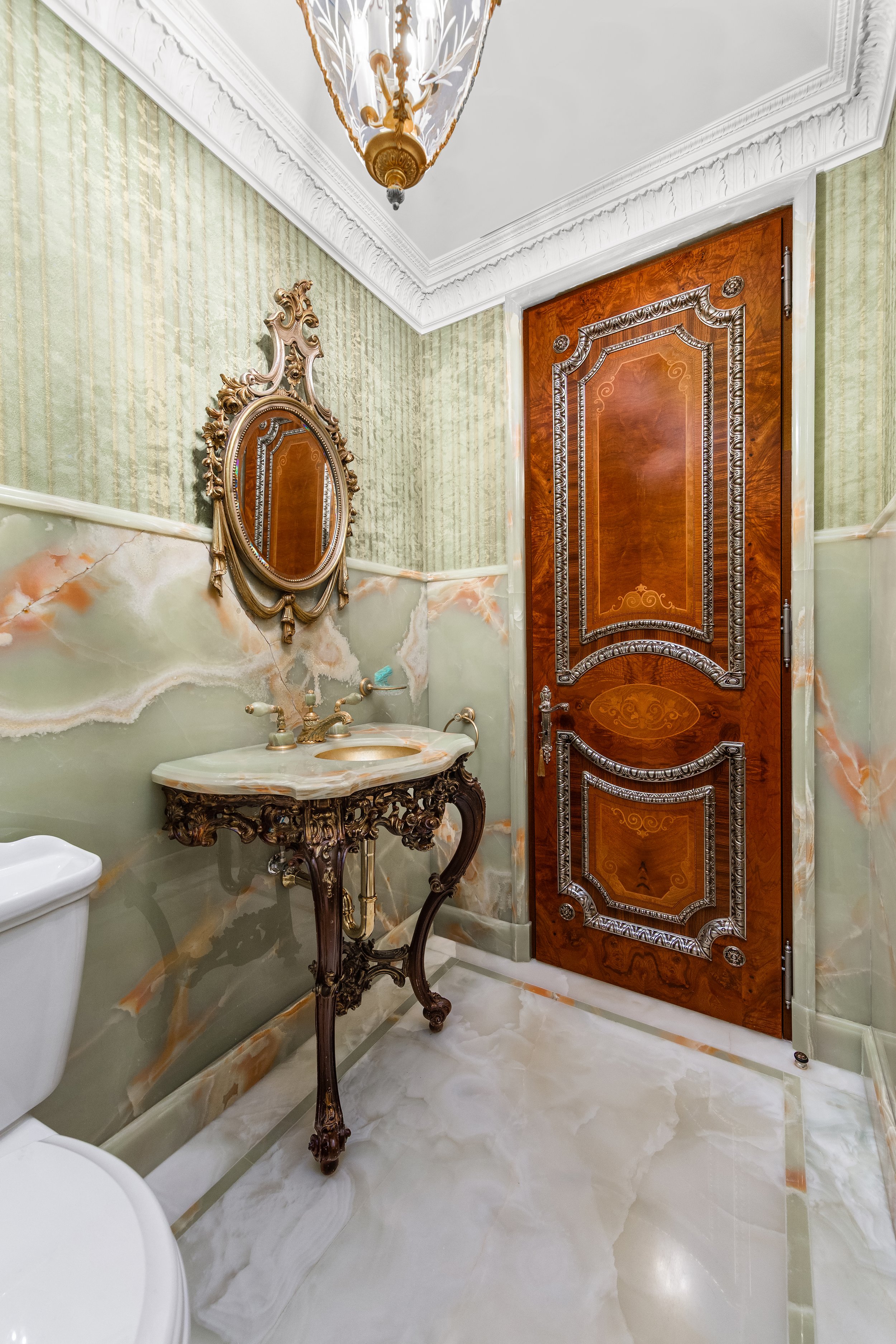 An ornate bathroom with green marble walls, a gold-framed mirror, a marble sink with decorative gold fixtures, a large wooden door with intricate silver inlay, and a white marble floor.