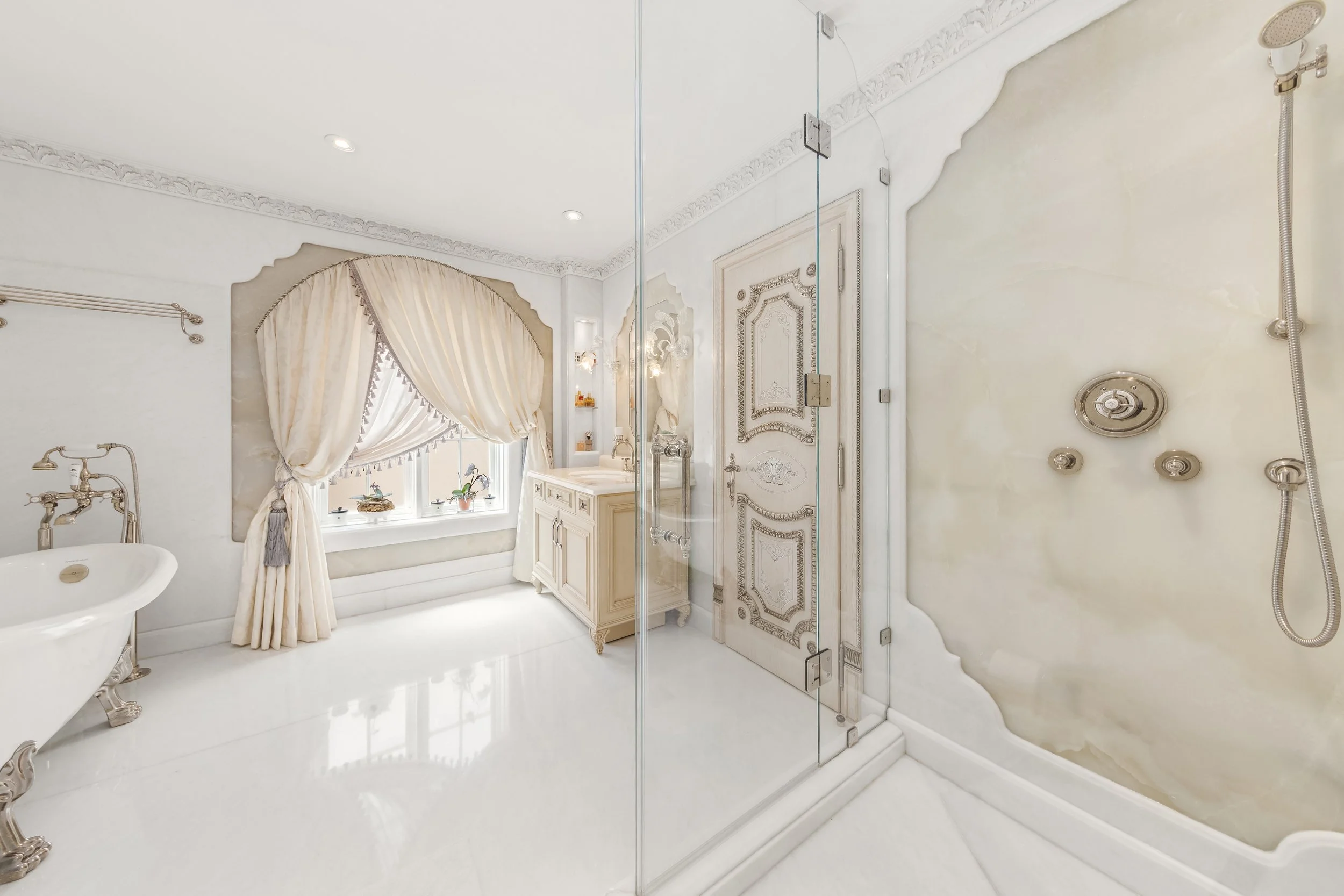 Luxurious bathroom with a glass shower enclosure, ornate door, vintage-style bathtub, large window with elegant curtains, cream-colored walls, and decorative crown molding.