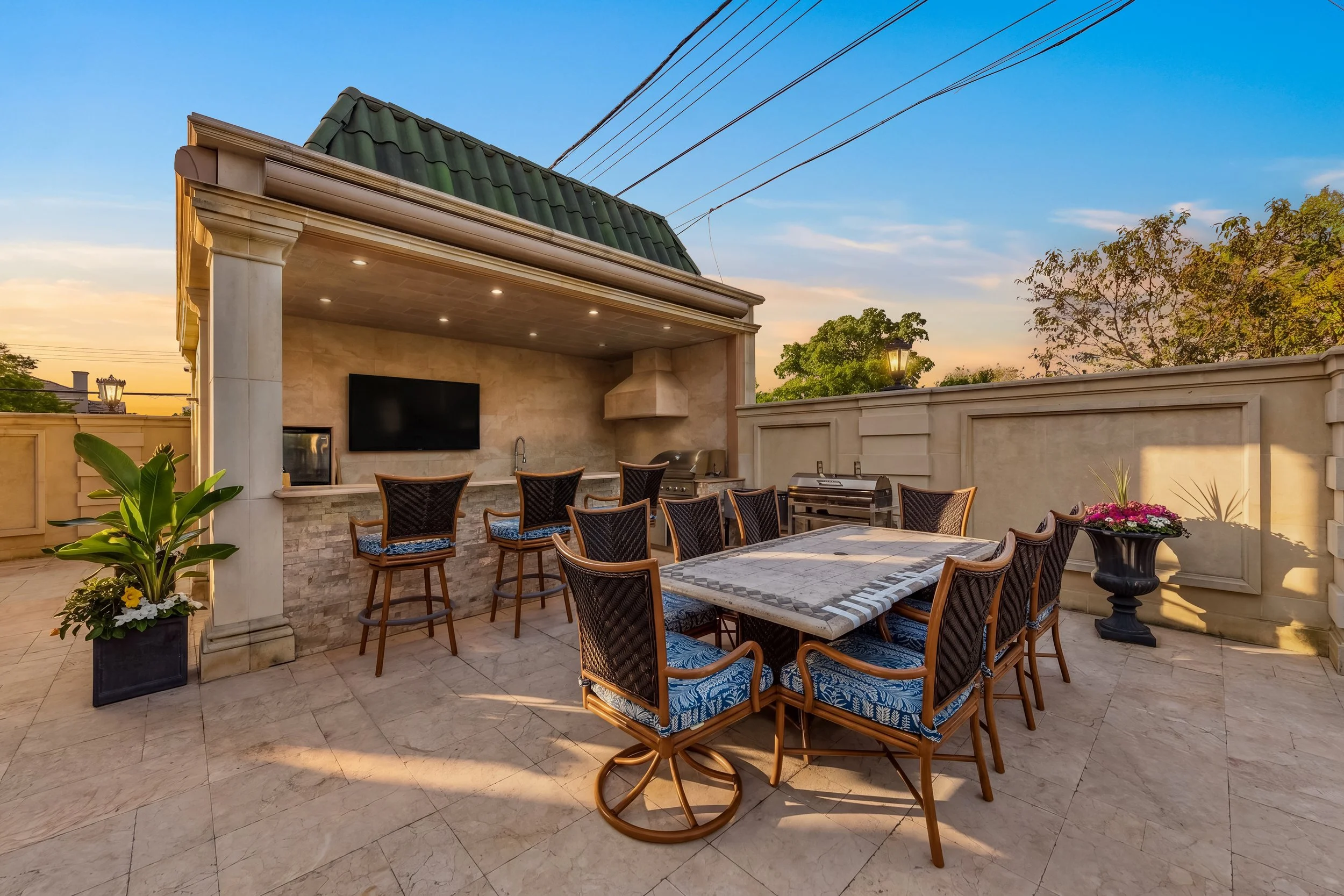 An outdoor patio with a dining table and chairs, a built-in outdoor kitchen with a TV, potted plants, and a stone fence with trees and a sunset sky in the background.