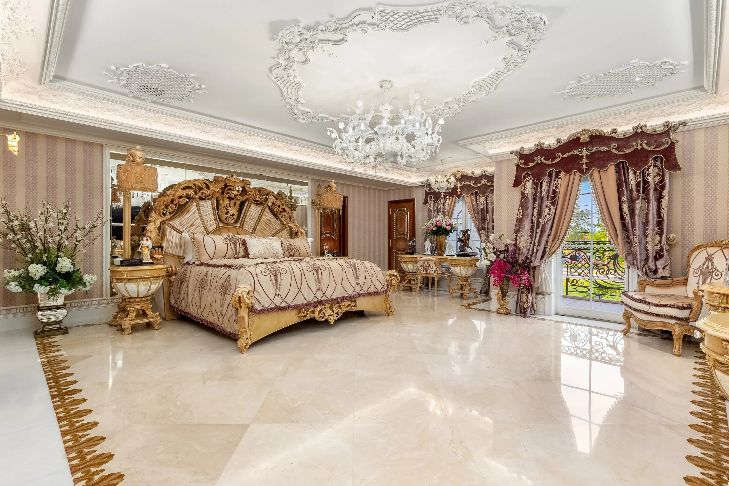 Luxurious bedroom with ornate gold-trimmed furniture, a large bed with decorative pillows, a chandelier, and elegant curtains.