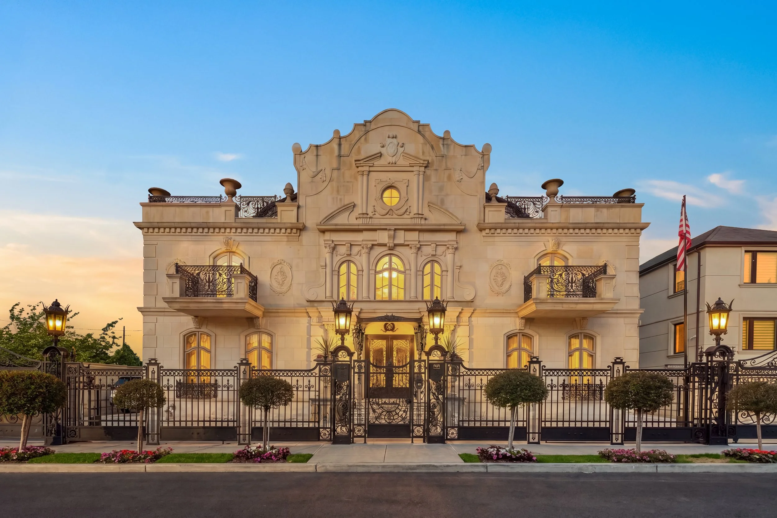 Elegant historic mansion with ornate architectural details, iron balconies, a gated front yard, and American flag on the side, illuminated by warm exterior lighting at sunset.
