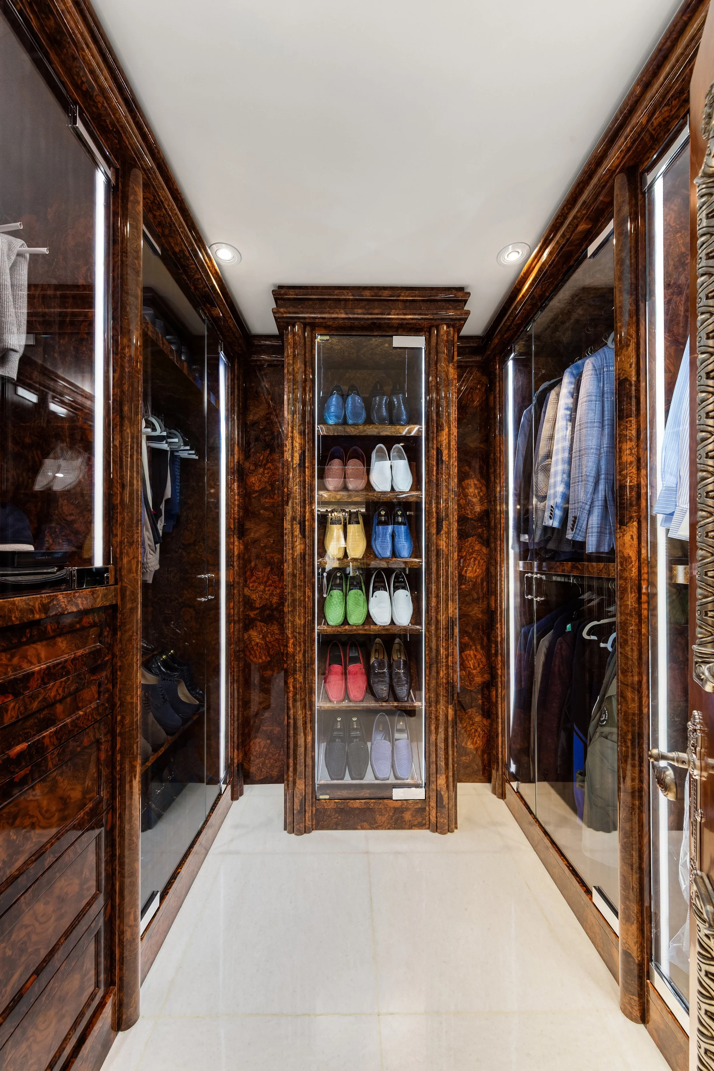 A walk-in closet with wooden shelves and glass doors filled with men's suits, shoes, and accessories.