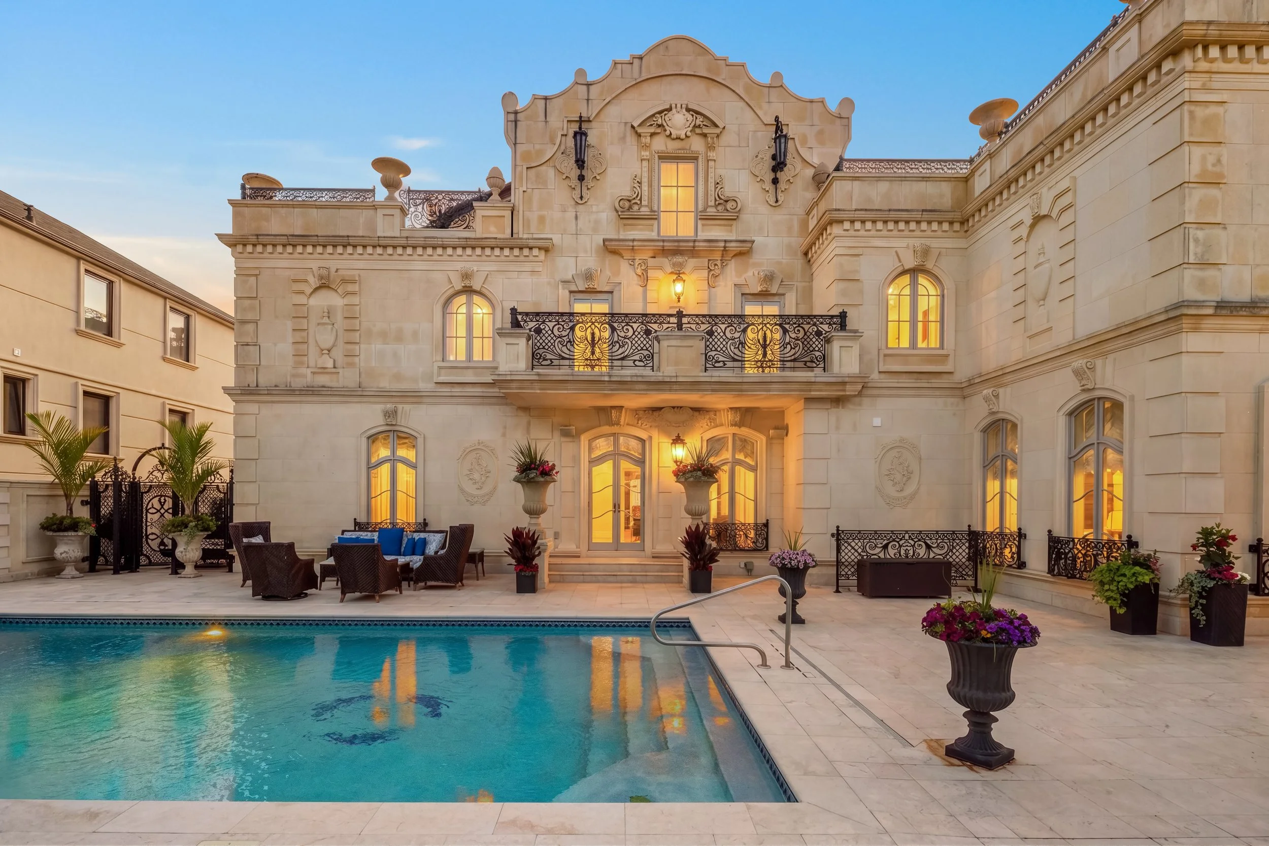 Luxury house with illuminated windows, a balcony, and a terrace area with potted plants, outdoor seating, and a swimming pool in the foreground.