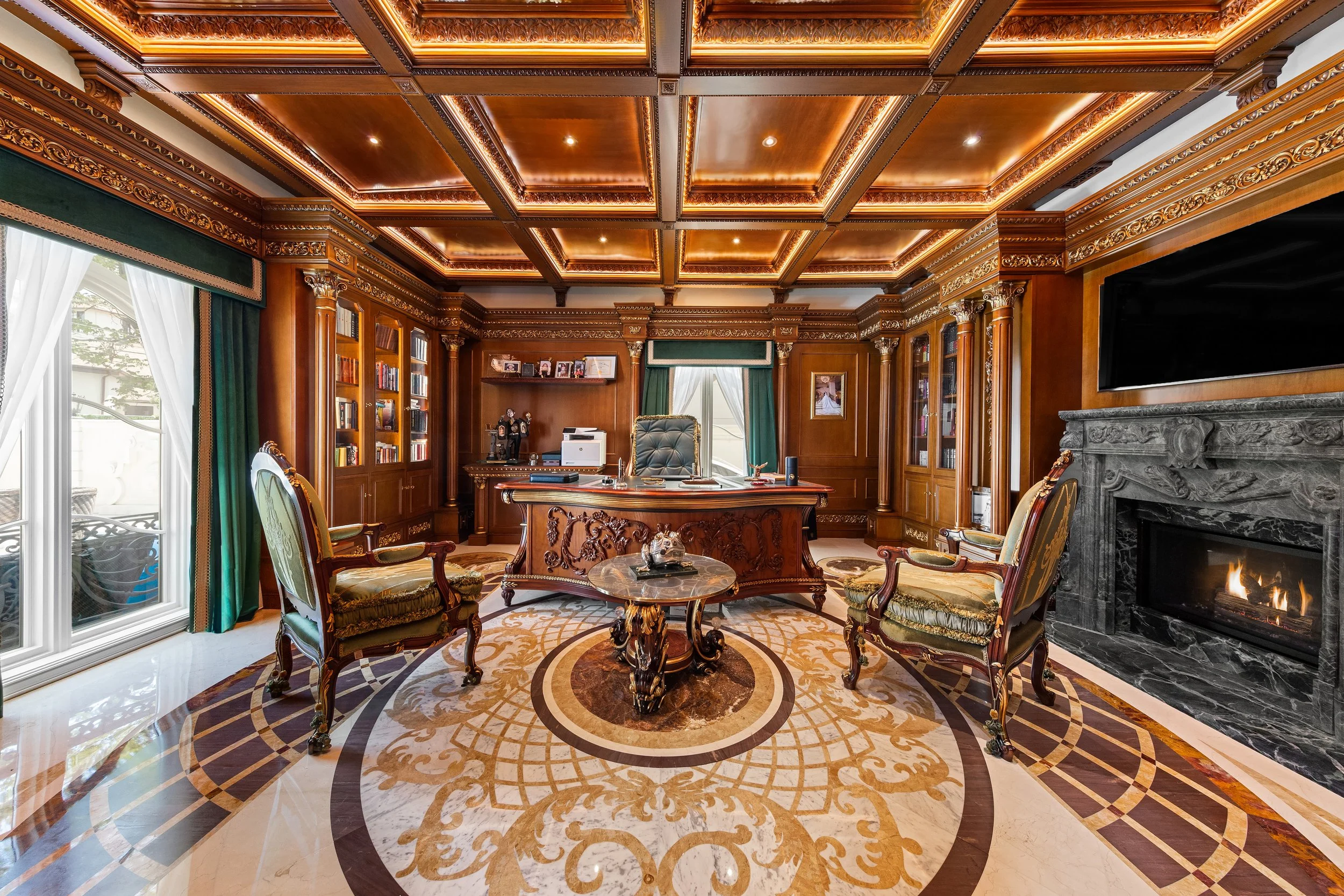 Luxurious wood-paneled office with coffered ceiling, ornate crown molding, and a large fireplace with a marble surround. Elegant chairs and a patterned area rug are in the room, with built-in bookshelves filled with books and decorative items. Large 