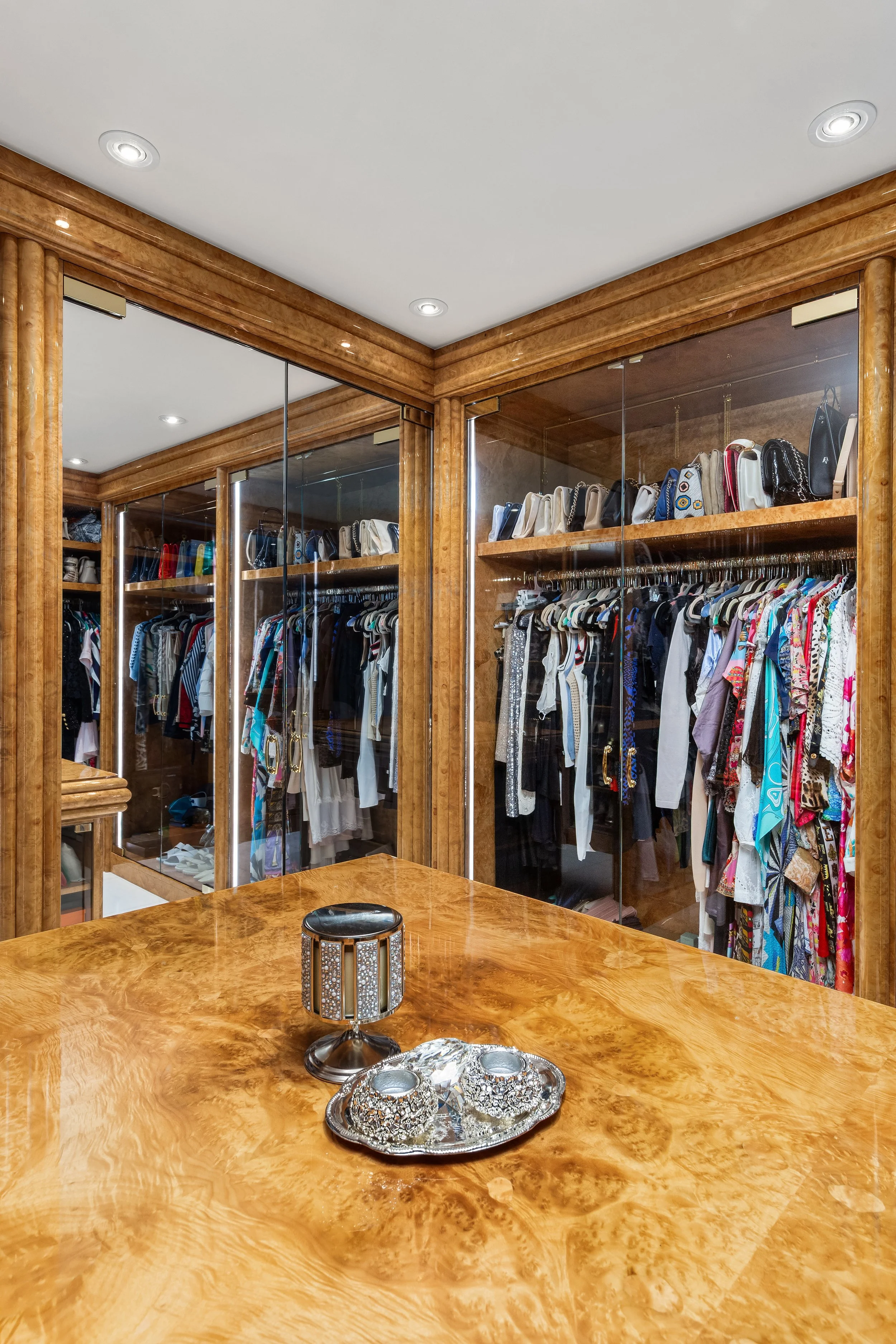 A walk-in closet with wooden shelves and racks filled with clothes and handbags, mirrored sliding doors, a wooden countertop, and decorative silver candles and a candle holder on the countertop.