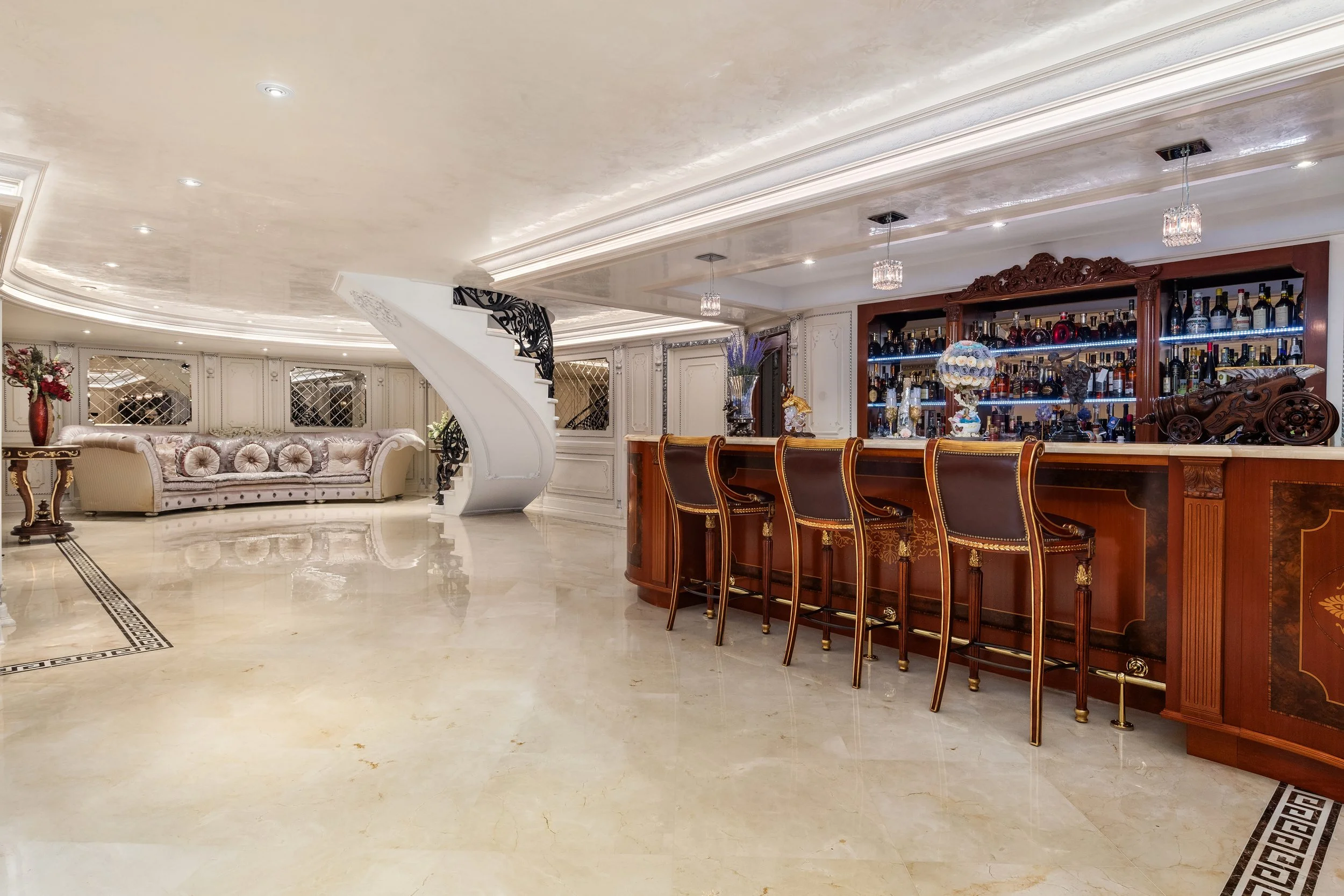 Luxurious living space with a curved staircase, ornate white and gold furniture, and a bar area with wooden bar stools, decorative bottles, and floral arrangements.