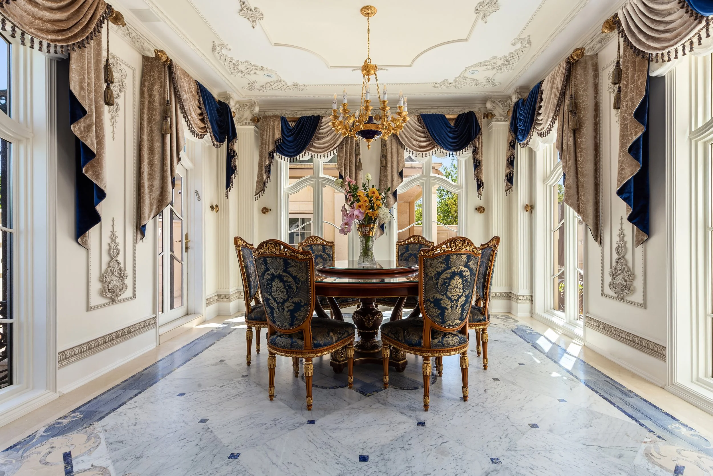 A luxurious dining room with large windows draped with ornate beige and blue curtains, a round wooden dining table with six upholstered chairs, a floral centerpiece, a gold and black chandelier, intricate white molding on the ceiling, and a marble fl