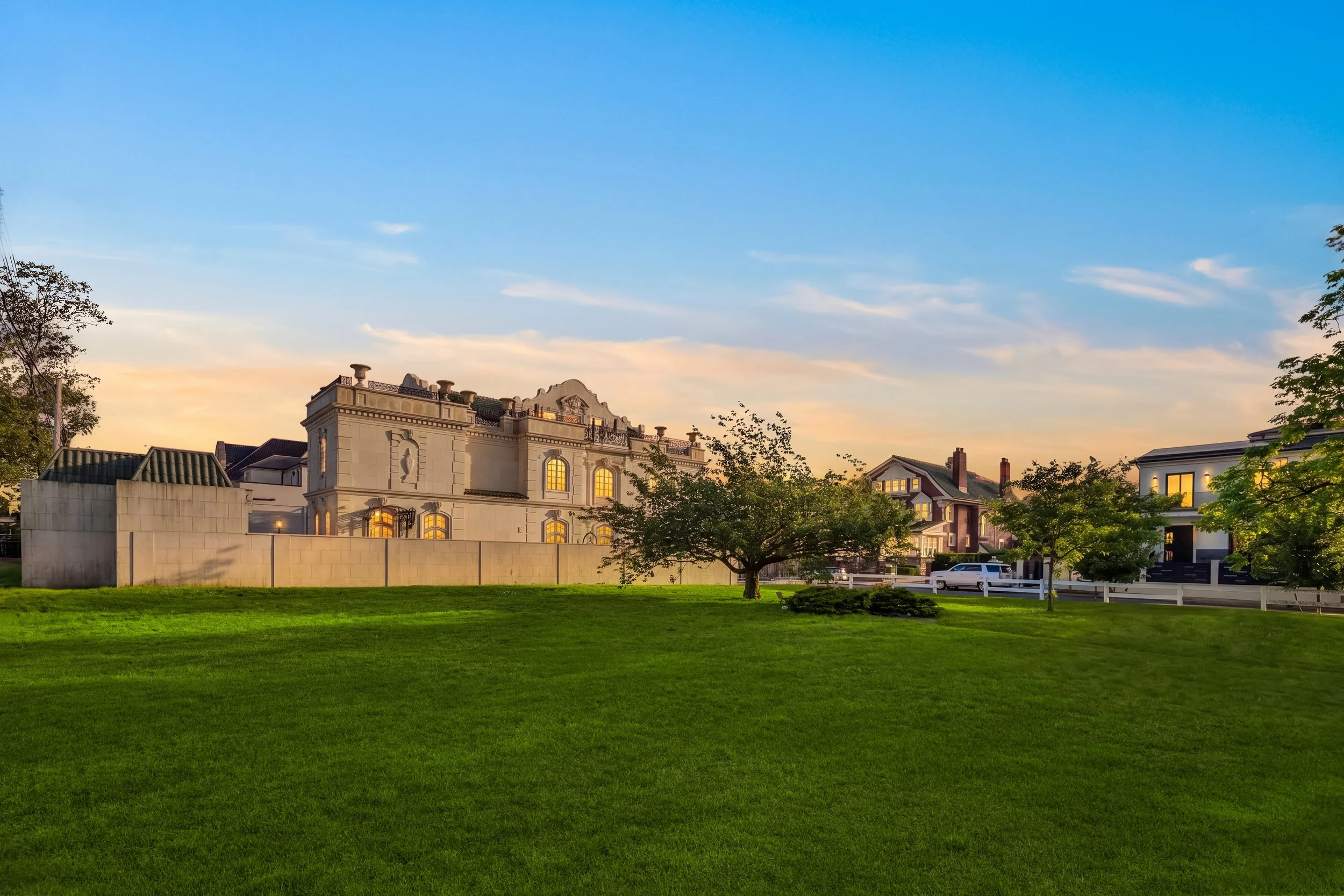 A residential neighborhood during sunset with a large green lawn, trees, and houses with lit windows, sky with clouds and a gradient from blue to peach.