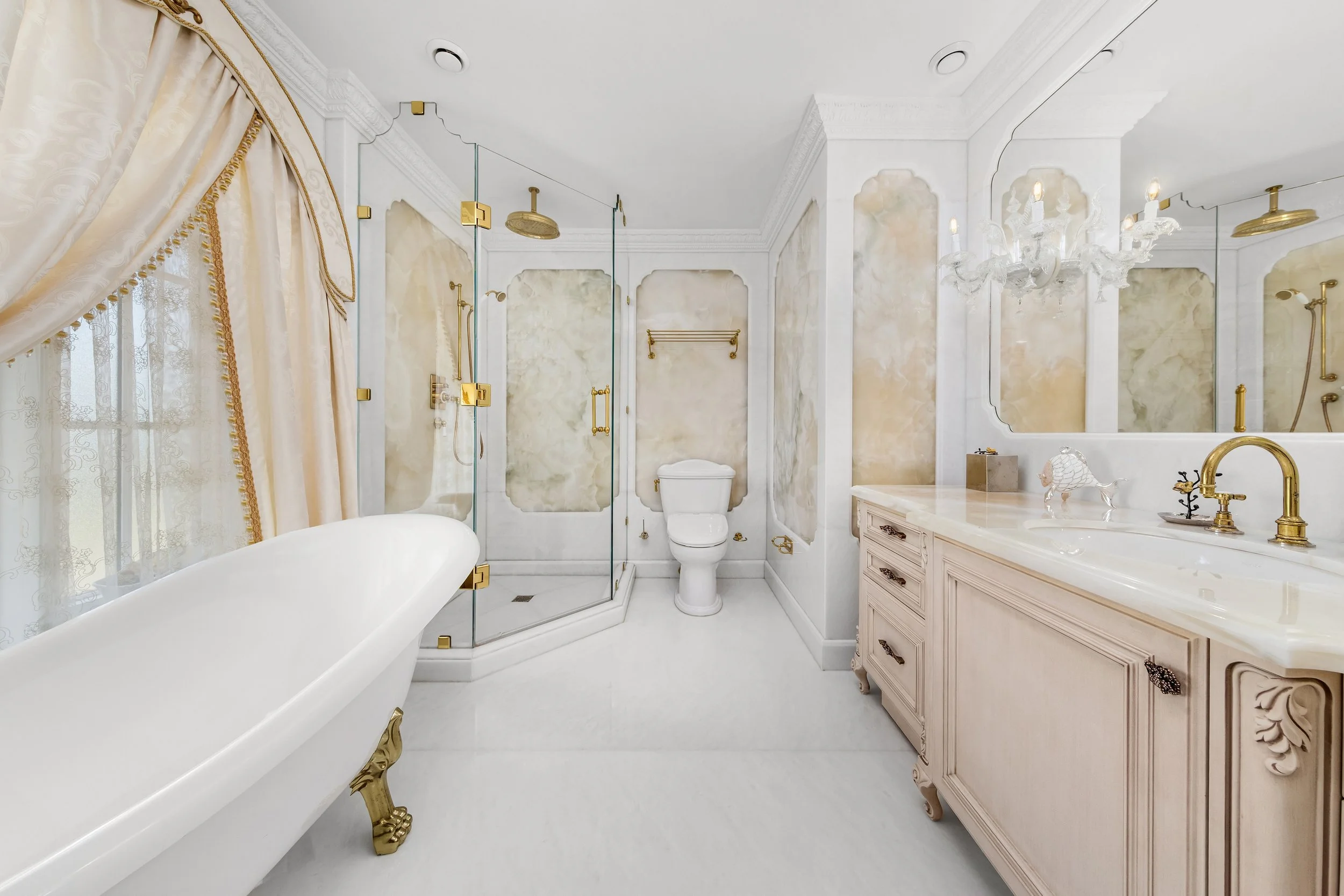 A luxurious bathroom featuring a freestanding clawfoot bathtub with gold accents, a marble vanity with a gold faucet, a glass-enclosed shower with gold fixtures, a toilet, ornate crown molding, a chandelier, and elegant drapery near a window.