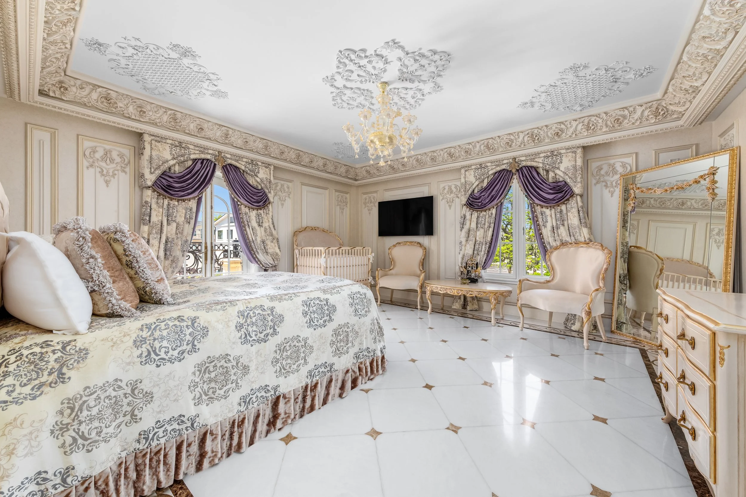 A luxurious bedroom with ornate white and gold furnishings, purple and cream drapes, a chandelier, a wall-mounted TV, a large mirror, and a polished marble floor.