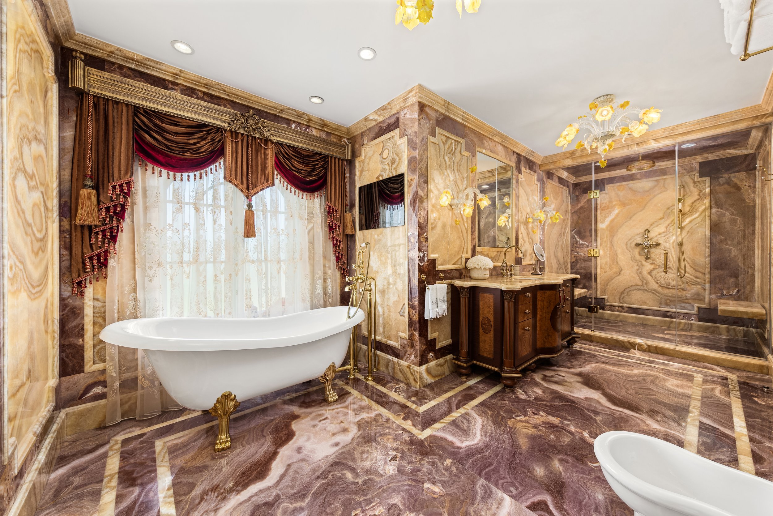 Luxurious bathroom with marble walls and flooring, a freestanding clawfoot bathtub with gold accents, a vanity with a mirror, a gold chandelier, and a glass-enclosed shower.