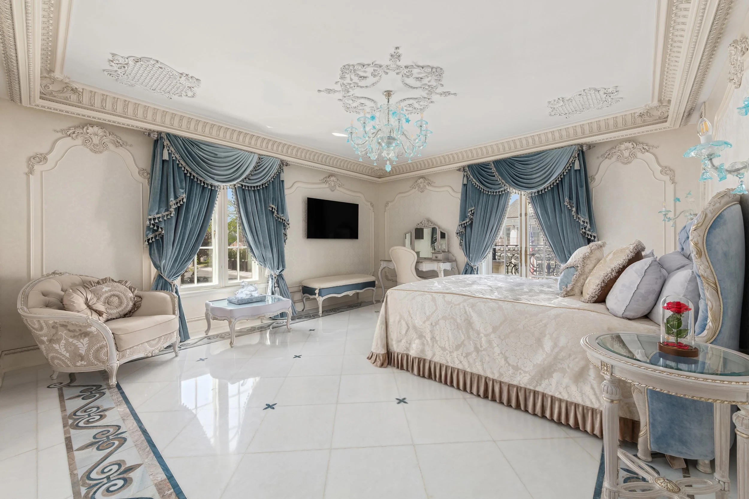 A luxurious bedroom with cream-colored walls, ornate crown molding, and a crystal chandelier. There are large blue curtains with tassels framing windows, and an elegant cream-colored bed with multiple decorative pillows. A vintage-style side table wi