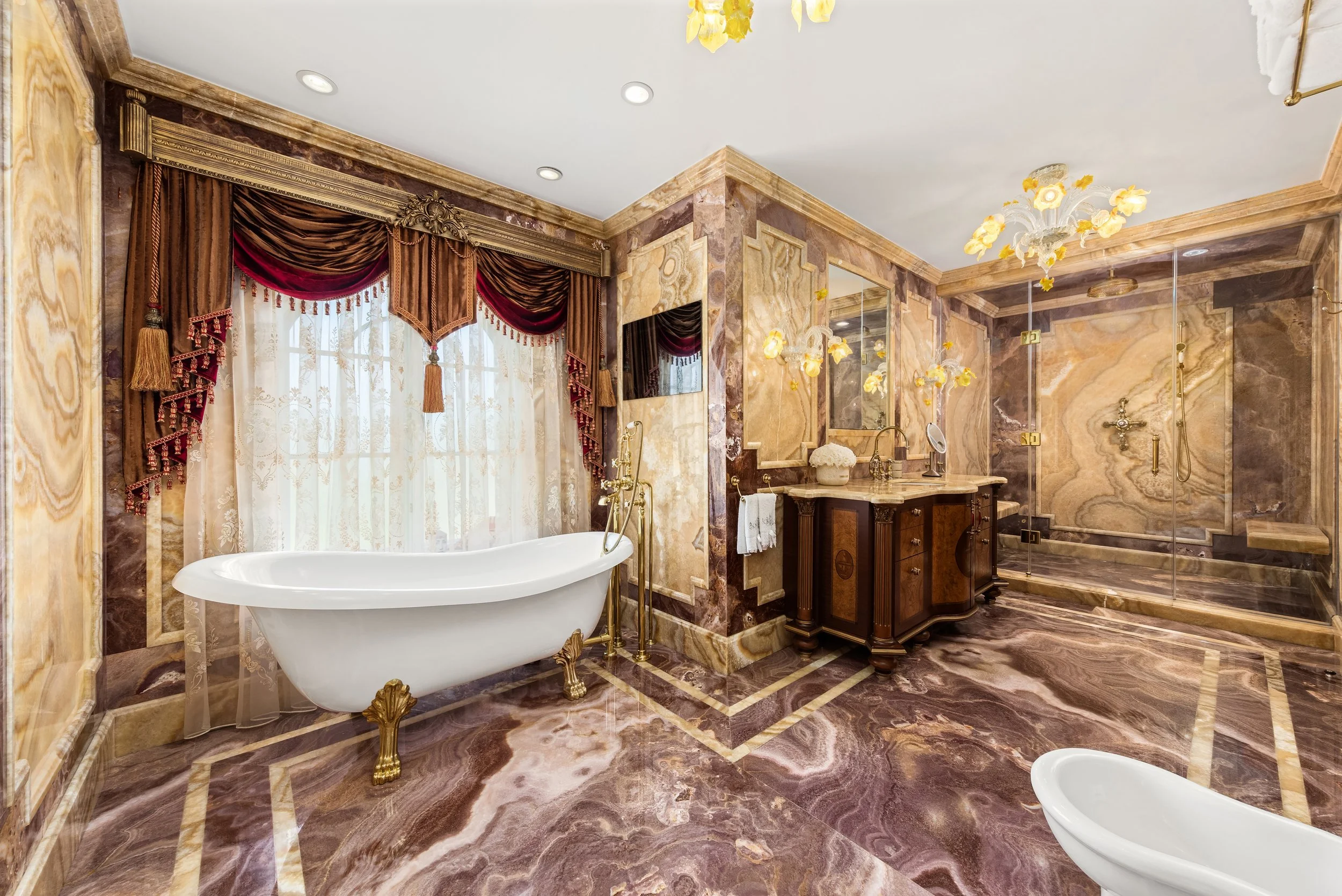 A luxurious bathroom featuring a clawfoot bathtub with gold fixtures, marble walls and floors, a large window with heavy draped curtains, a wooden vanity with a marble countertop, and a separate shower stall with glass doors, ornate chandeliers, and 