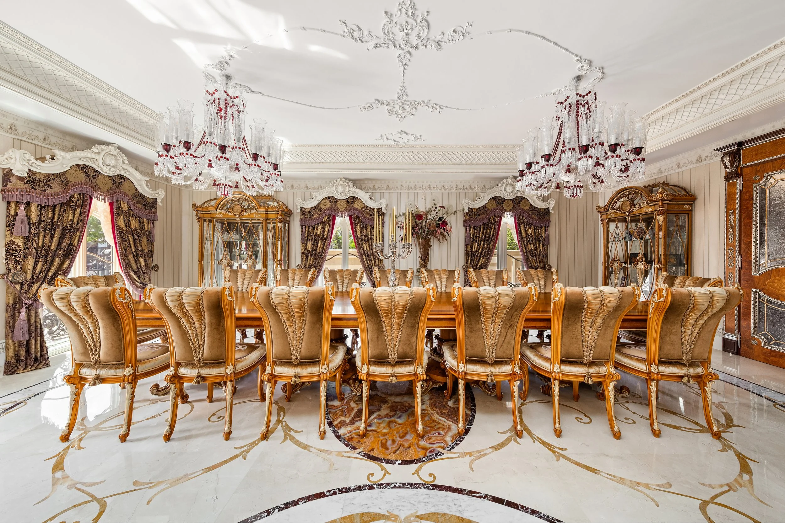 A luxurious dining room with a long wooden table surrounded by ornate, high-backed chairs. The room features elegant chandeliers hanging from a decorated ceiling, pastel striped wallpaper, and ornate curtains with gold and purple patterns framing lar