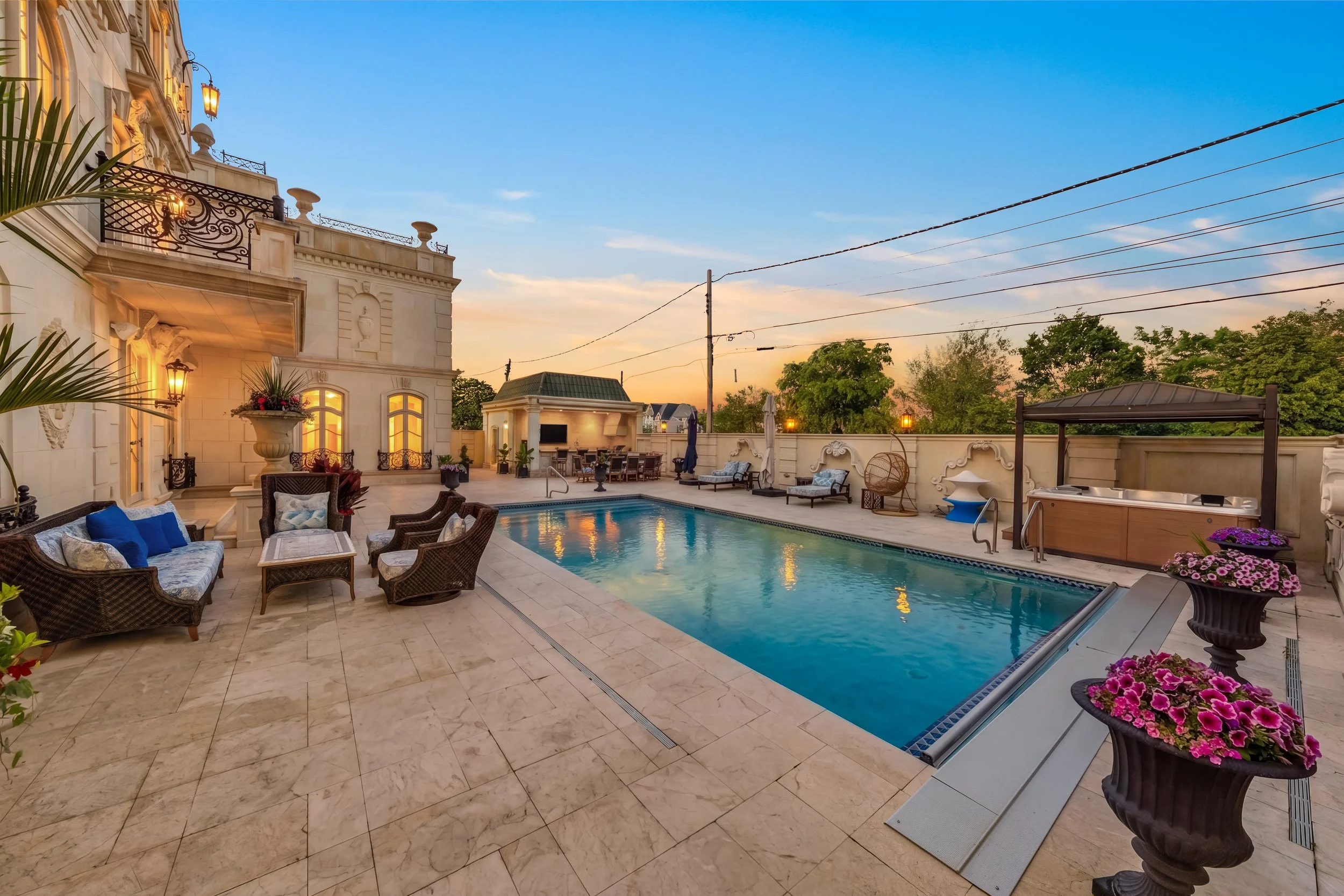 Luxury backyard with swimming pool, outdoor seating, potted flowers, hot tub, and sunset sky.