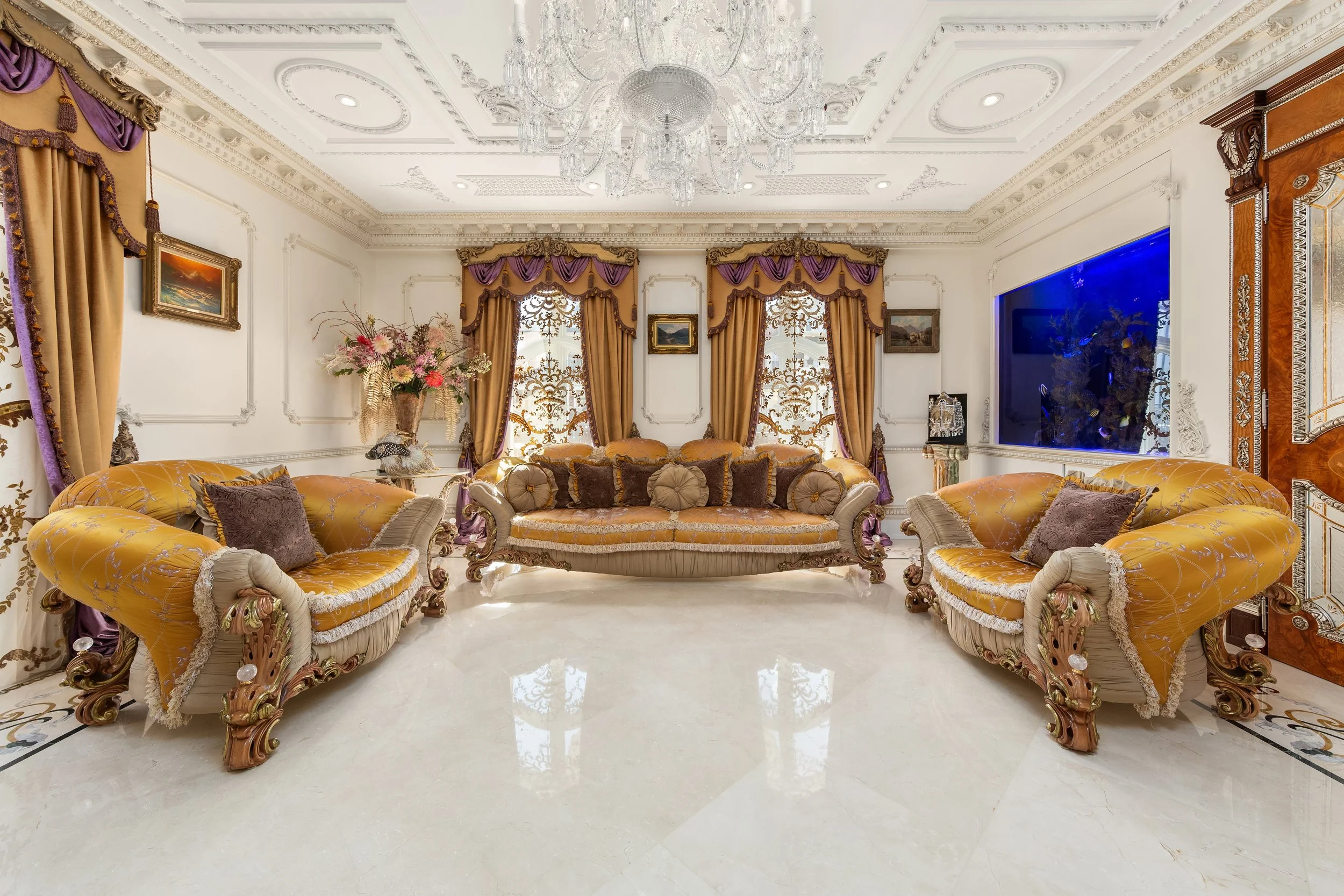 Luxurious living room with ornate gold and purple couches, gold curtains with purple accents, a large floral arrangement, a chandelier, and a large fish tank on the wall.